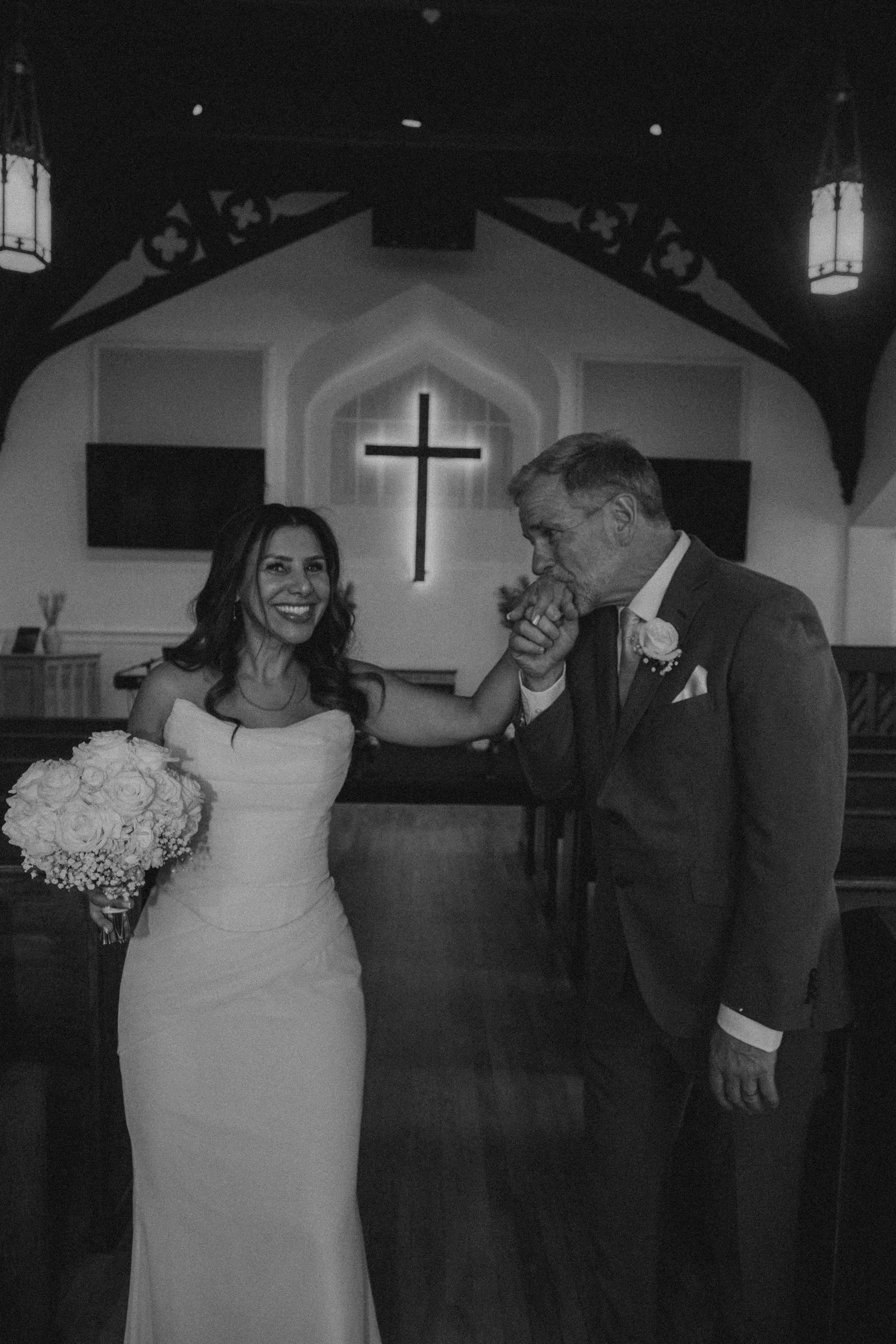A bride in a white wedding gown holding a bouquet of flowers, dancing with an older man in a suit at a wedding reception inside a church with a cross in the background.
