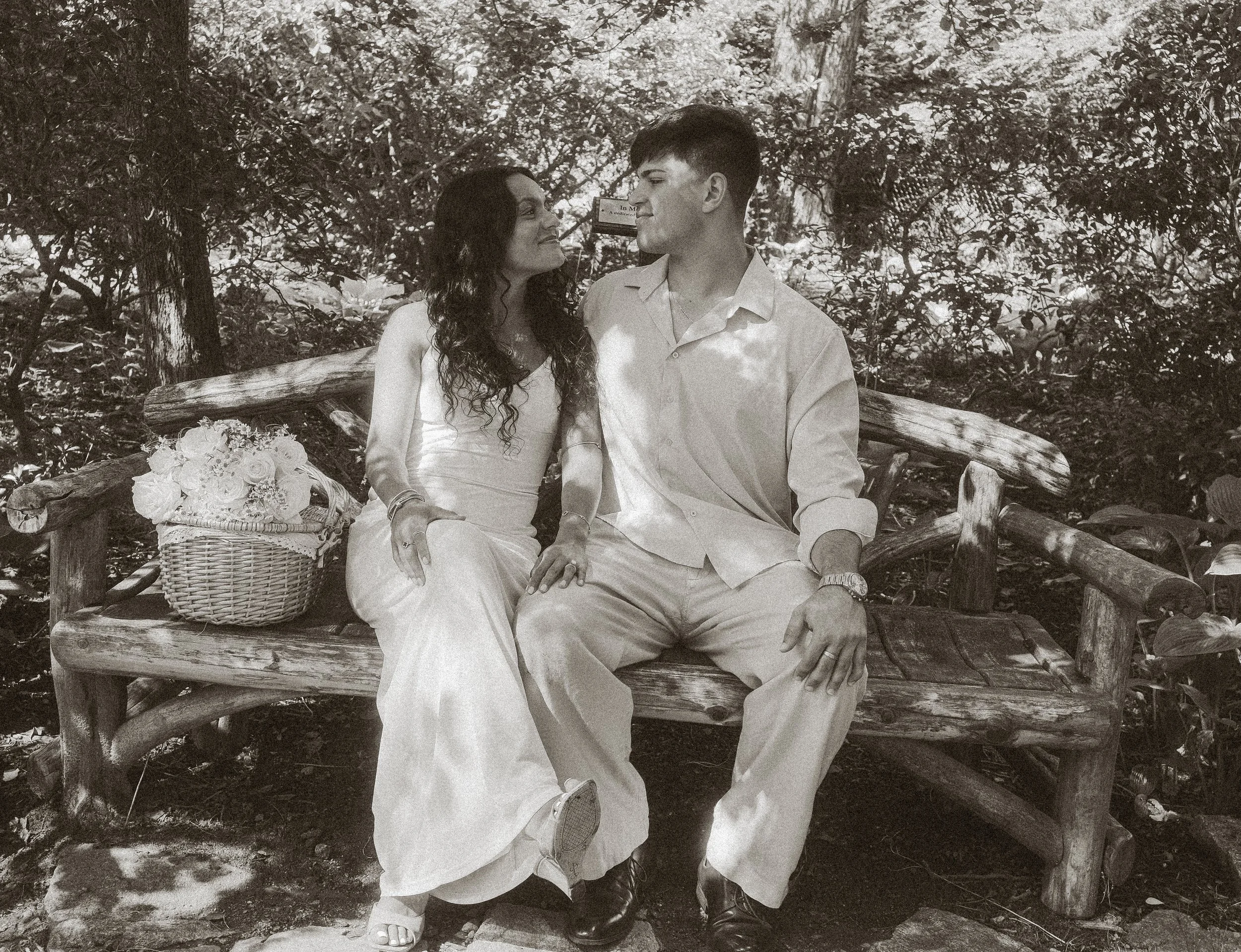 A couple sitting on a rustic wooden bench outdoors, with trees and foliage in the background. The woman is wearing a light-colored sleeveless dress and holding a bouquet, while the man, dressed in a light shirt and trousers, is sitting close to her, 