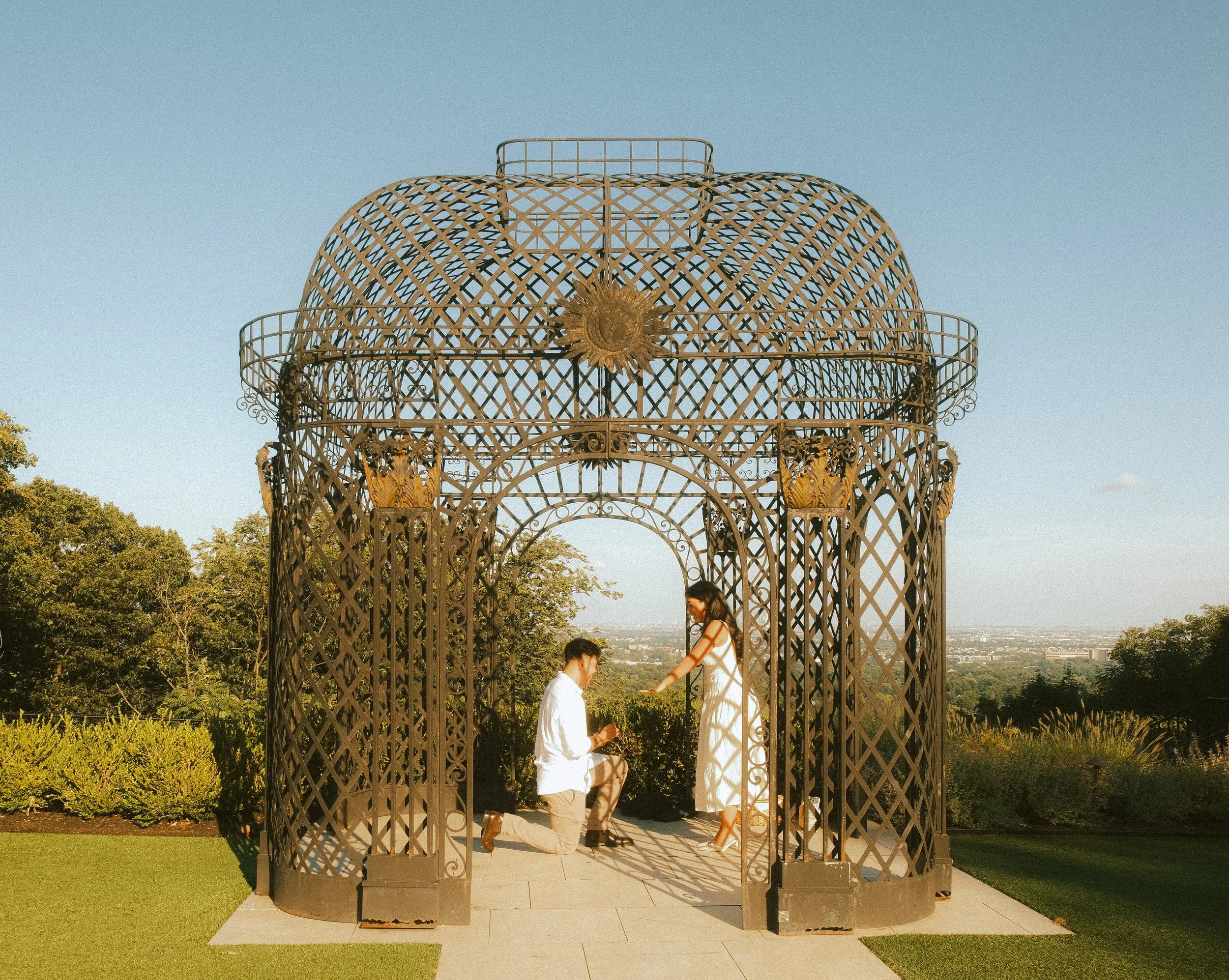 A man kneeling and a woman standing inside a decorative metal gazebo with a golden emblem on top, overlooking a cityscape and trees on a clear day.