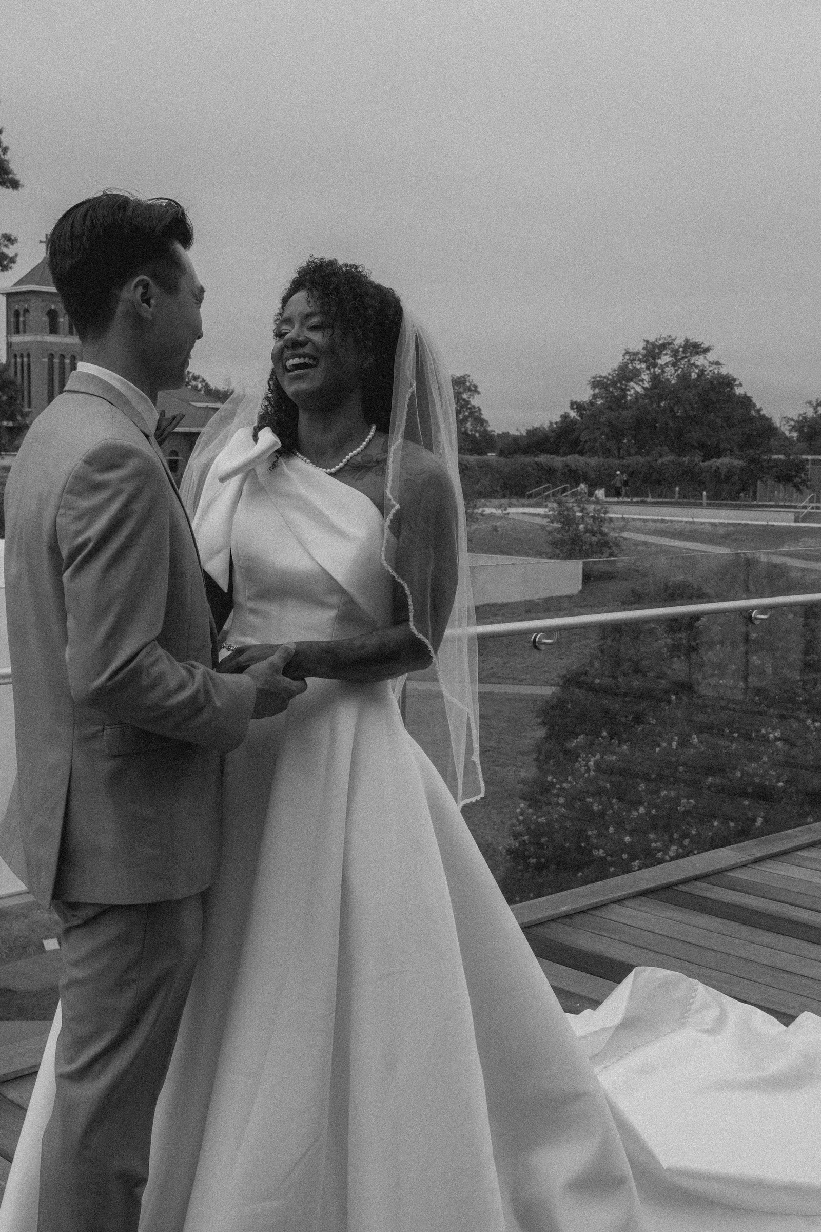 A bride and groom smiling and looking at each other on their wedding day, outdoors with a church and trees in the background.