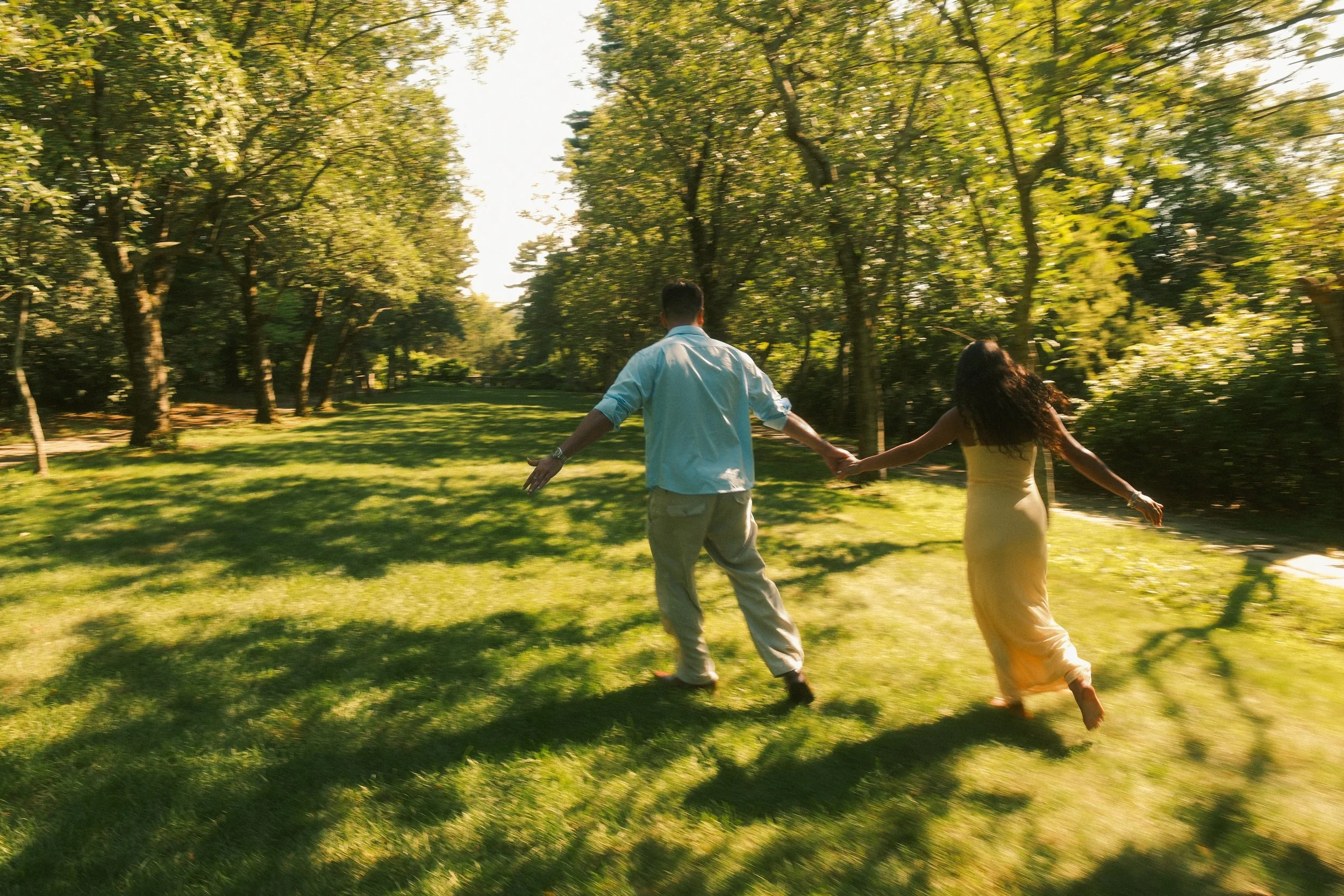 A couple is holding hands and spinning in a green park with trees and sunlight.