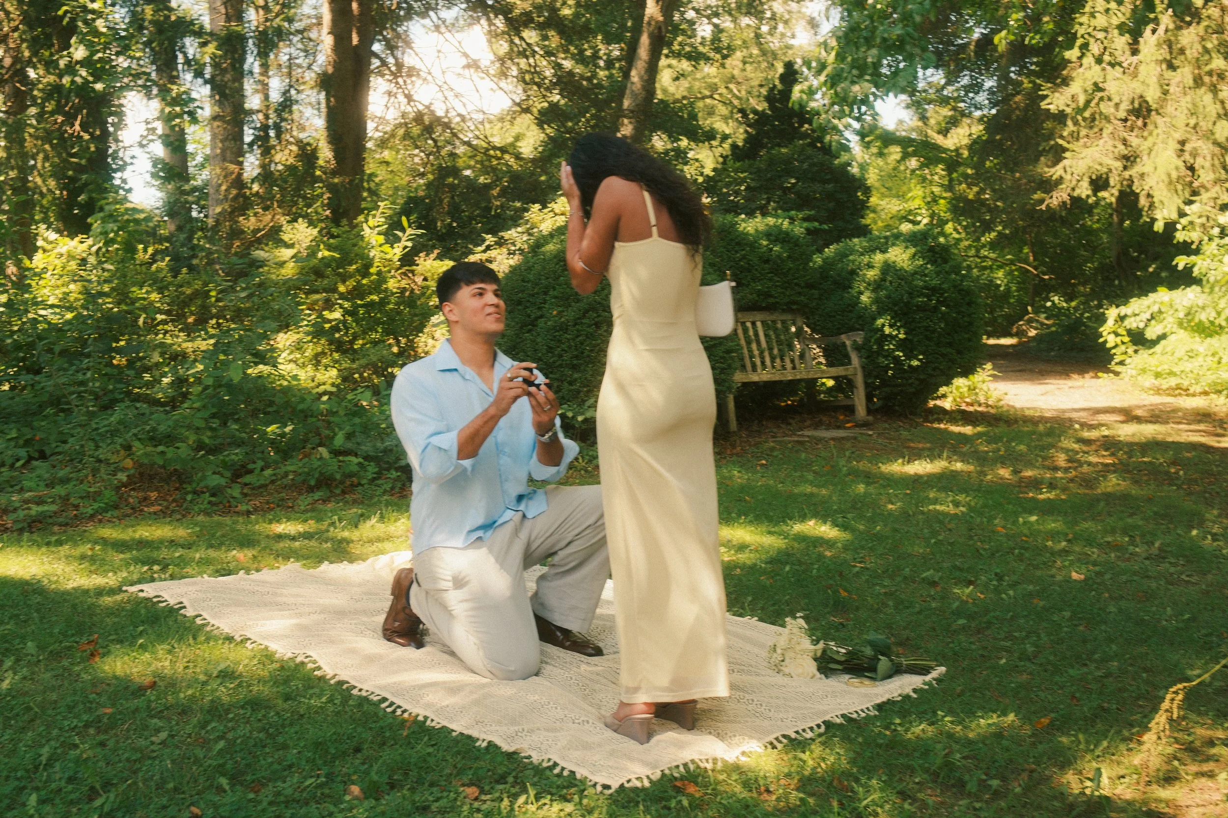 A man proposes marriage to a woman in a wooded park, with the man kneeling on one knee holding a small box and the woman covering her face in surprise or emotion.