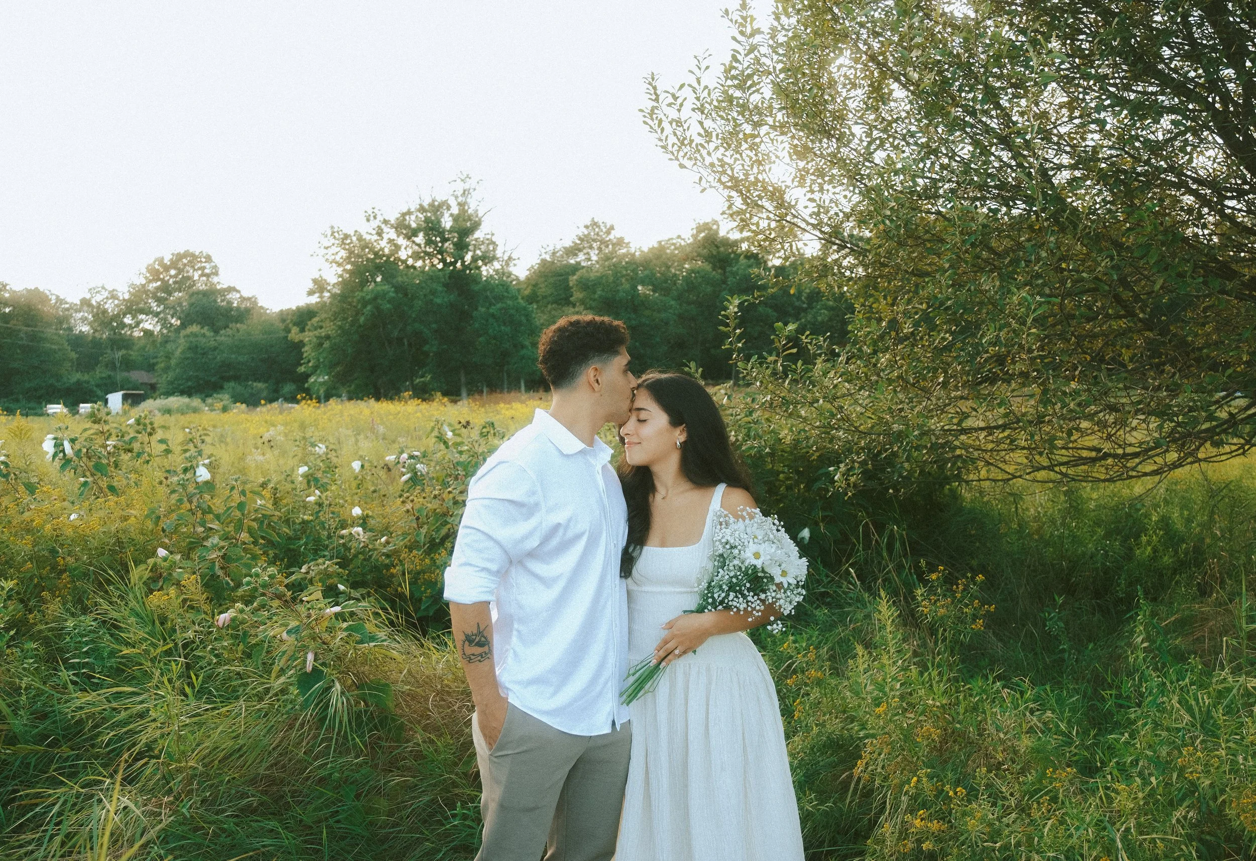 A couple stands in a field with lush greenery and yellow flowers, the man is kissing the woman's forehead, she is holding a bouquet of white daisies, and they both appear happy and serene.