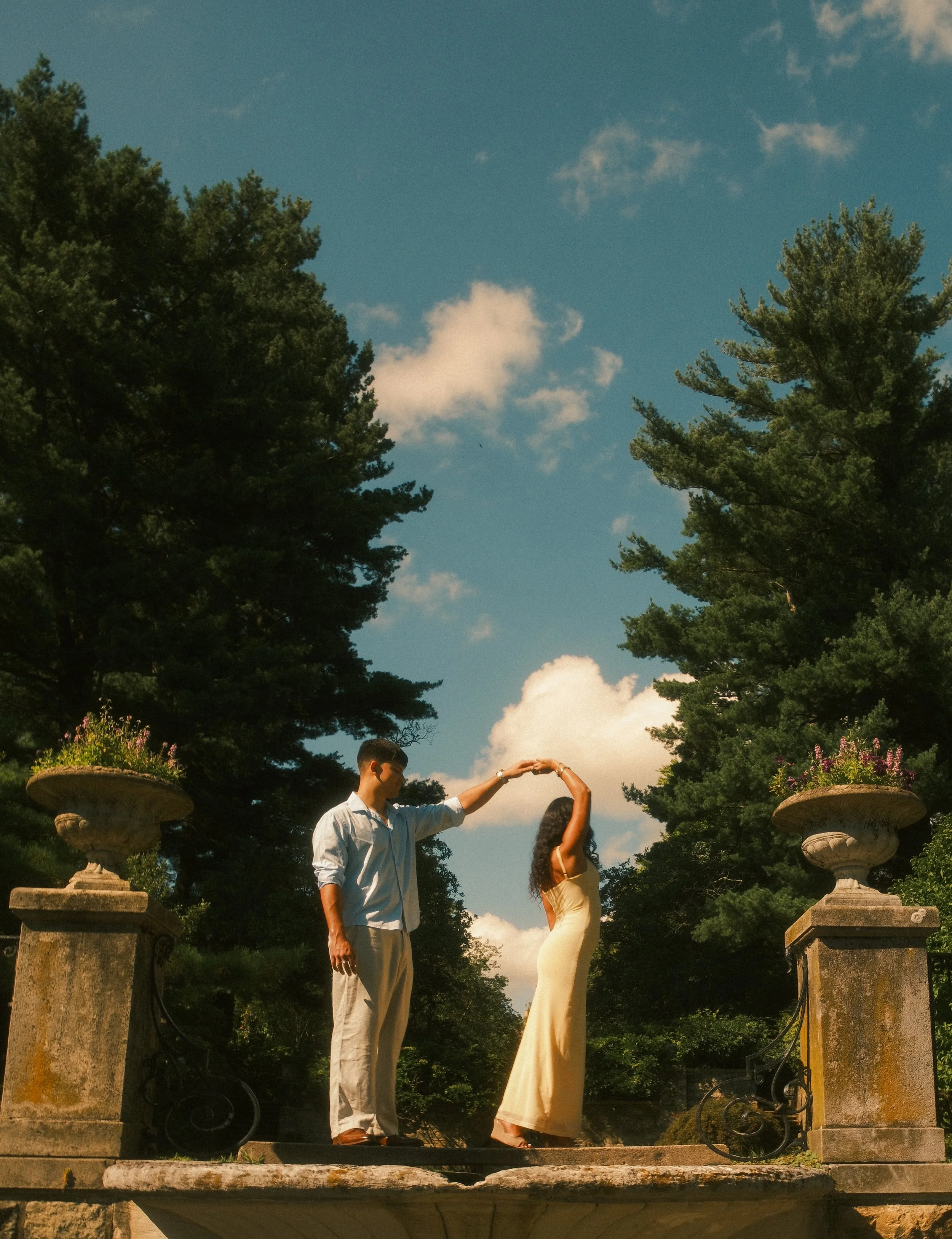 A man and woman holding hands and dancing outdoors on a stone balcony with trees and a blue sky with clouds in the background.