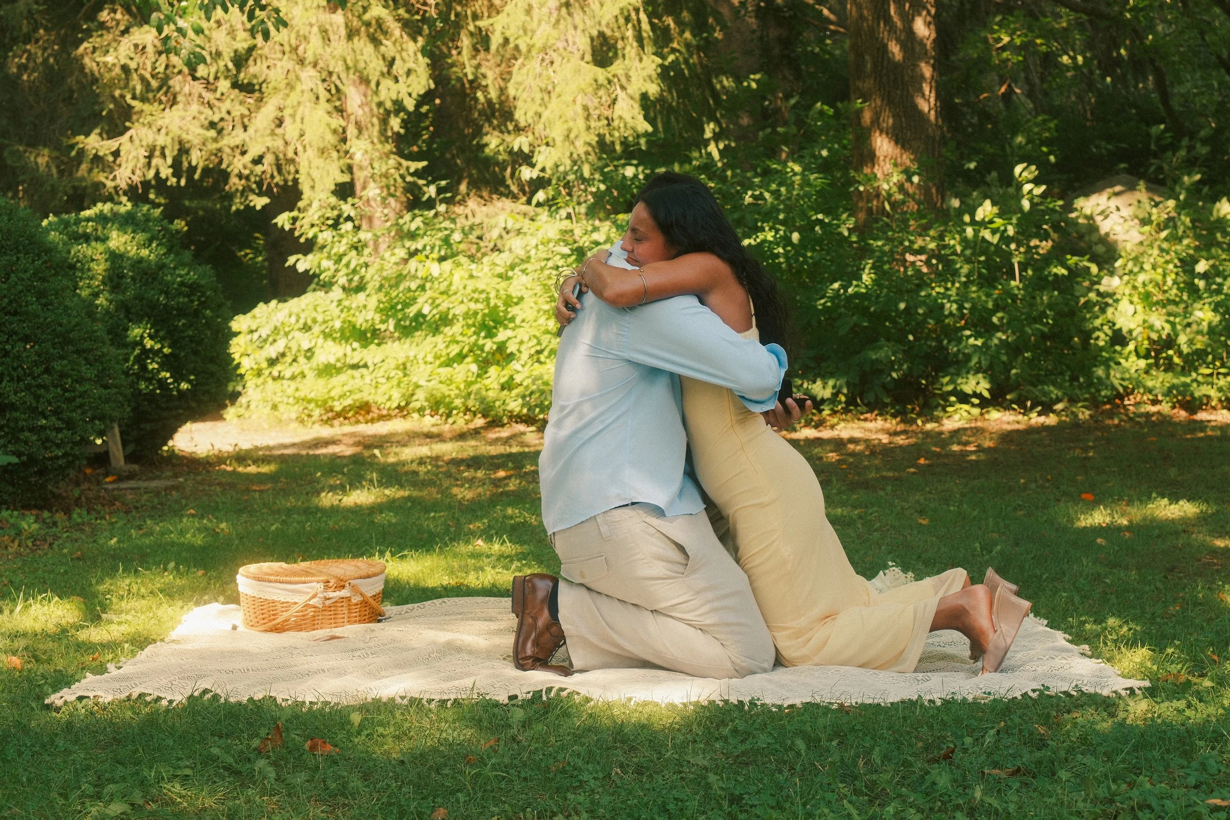 A woman and a man sharing a hug outdoors on a green lawn surrounded by trees, with a picnic basket nearby.