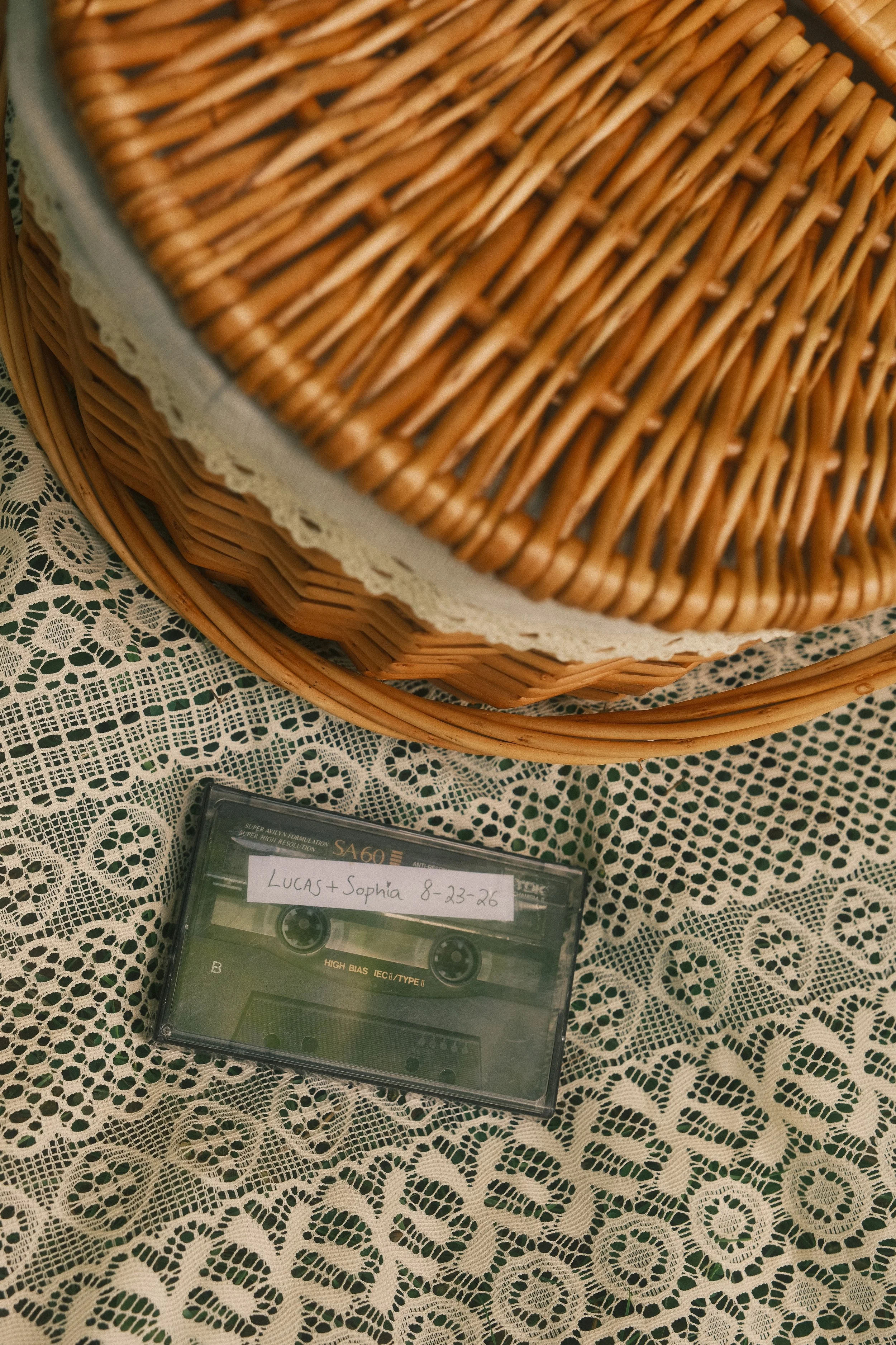 A woven wicker basket lined with white lace, placed on a lace tablecloth. In front of the basket, there is a cassette tape case labeled with the handwritten names 'LUCAS + SOPHIA 8-23-26'.