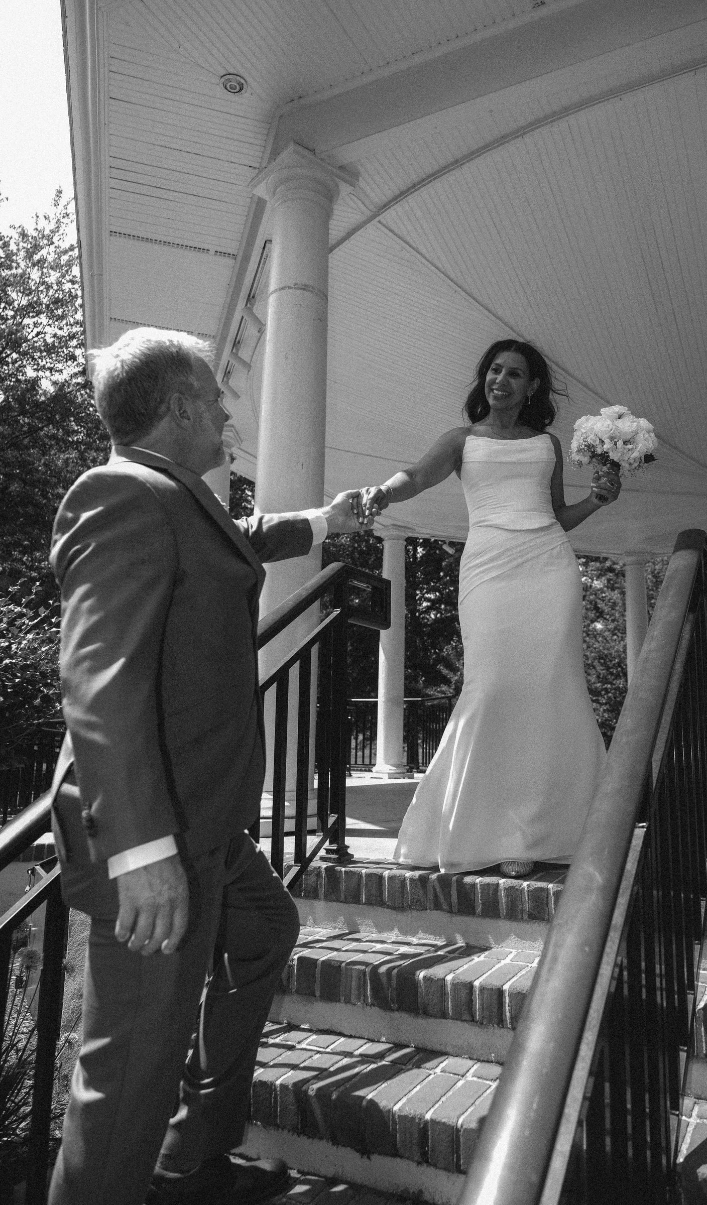 A woman in a wedding dress holding a bouquet greeting a man in a suit on the steps of a house or venue, with a porch and columns in the background.