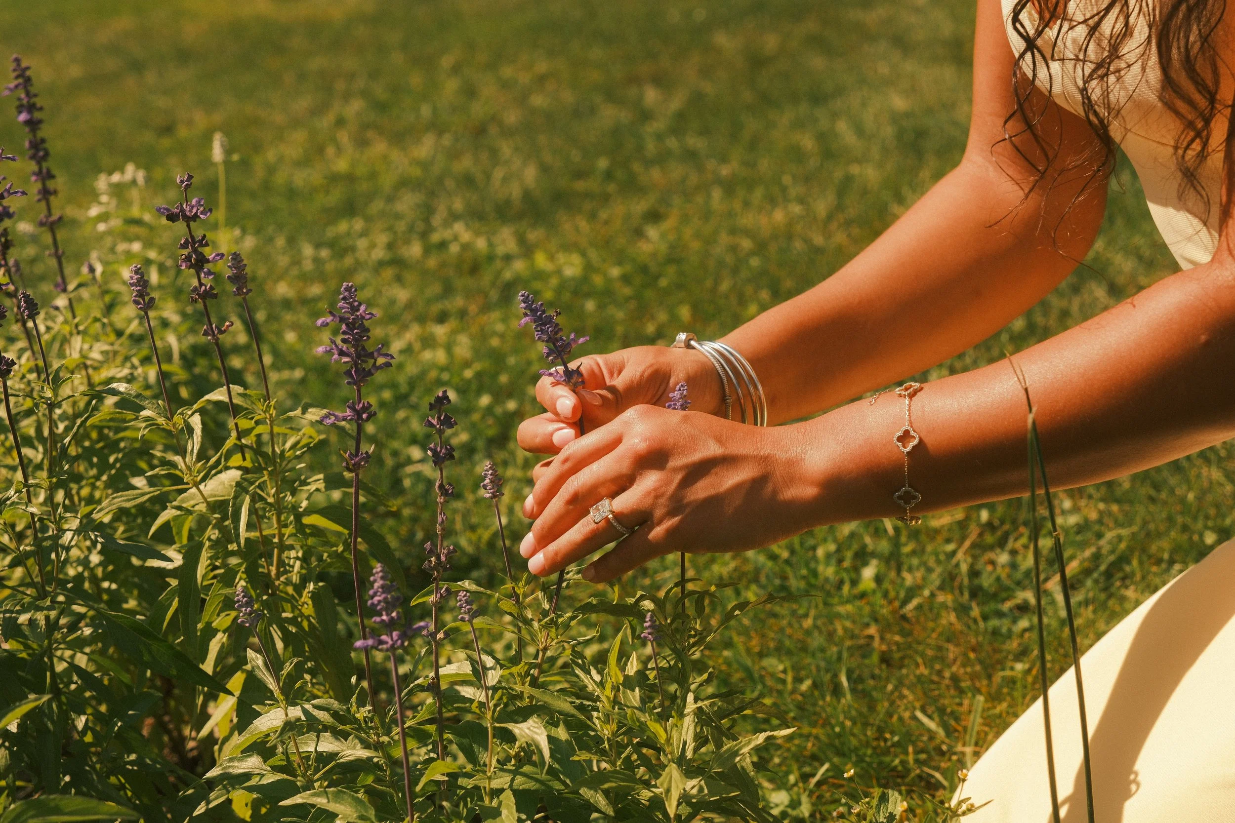 A woman with long curly hair is kneeling in a garden, holding a purple flower among green plants.