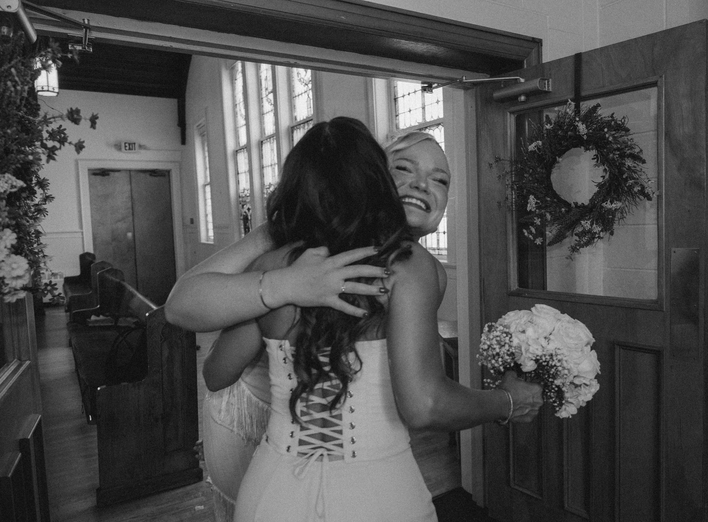 A joyful woman with platinum blonde hair smiling and hugging another person in a church or chapel decorated for a special occasion, possibly a wedding, with floral arrangements and a large wreath on the wall behind her.