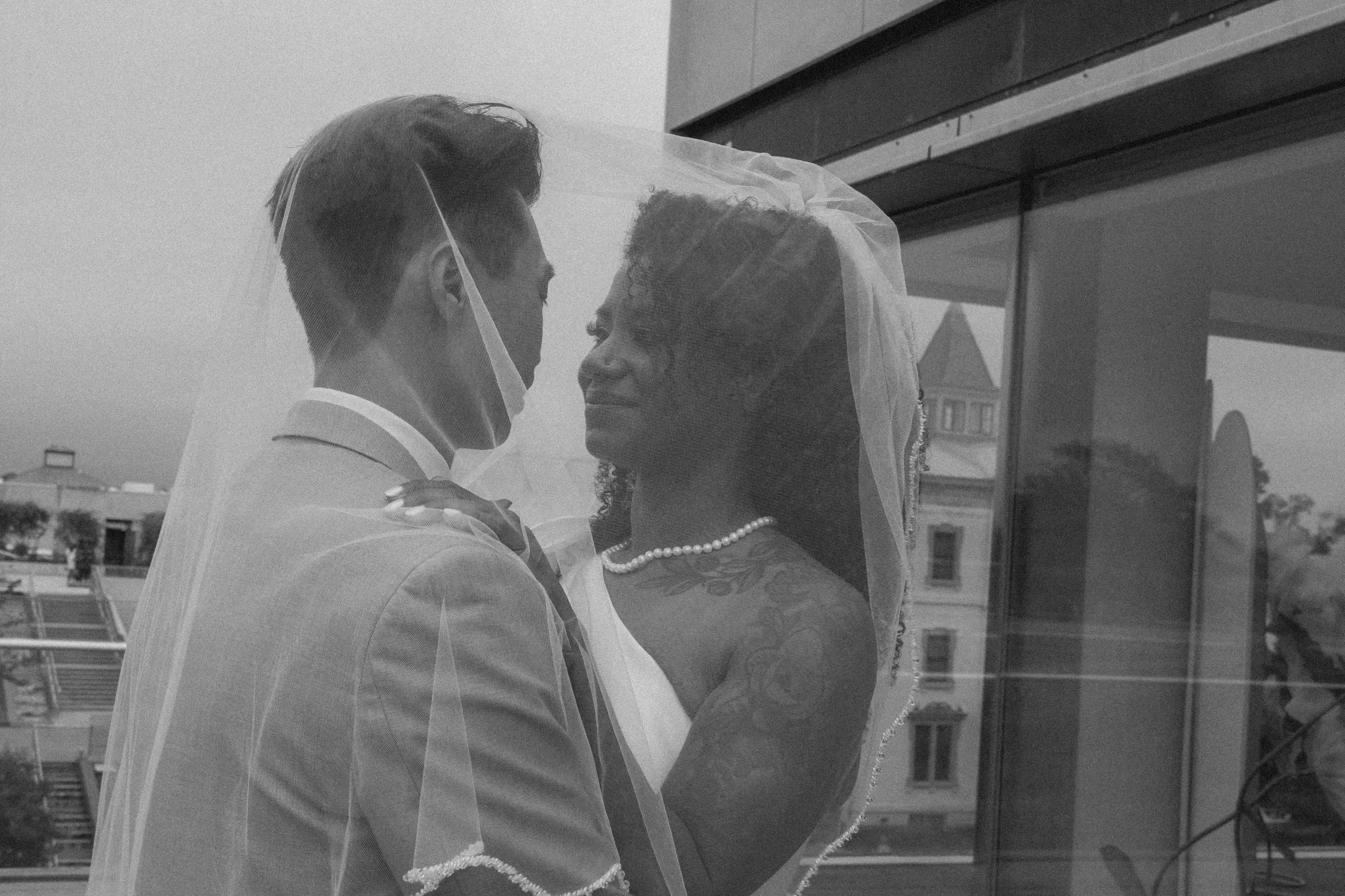 A black-and-white photo of a bride and groom with their foreheads touching, inside a building with cityscape visible through a window.