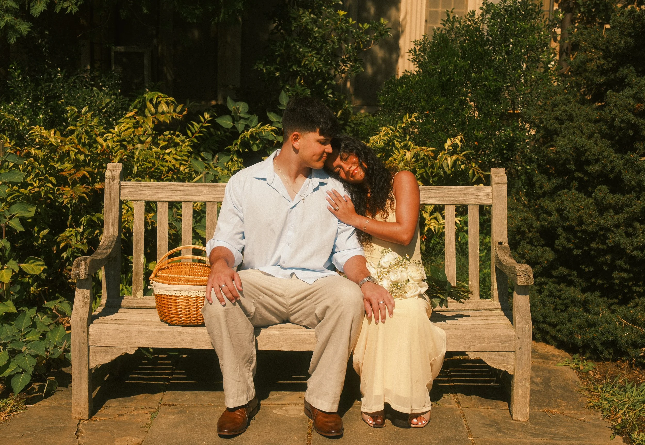 A couple sitting on a wooden bench outdoors surrounded by greenery, with the woman leaning her head on the man's shoulder, both smiling.