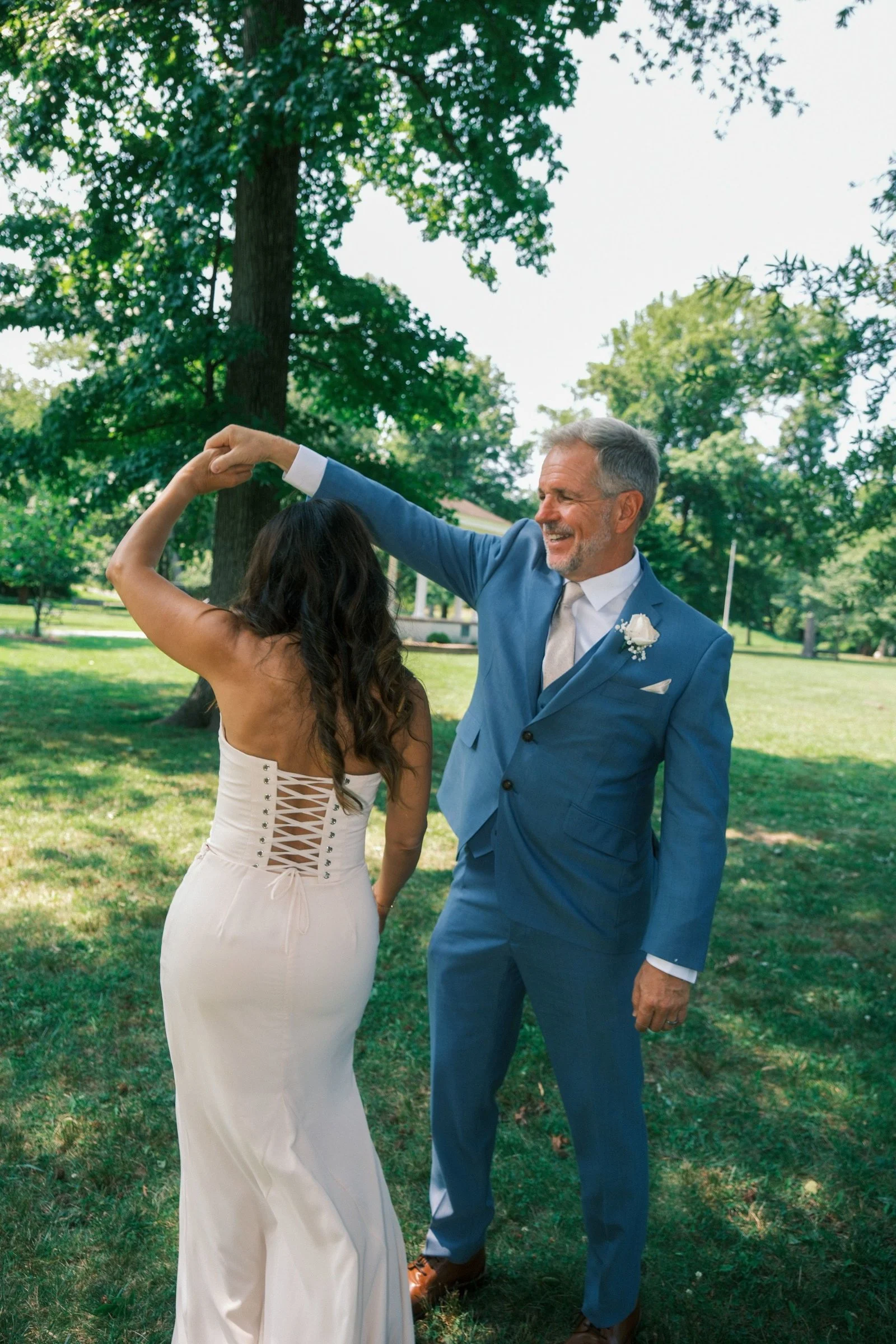 An older man in a blue suit dancing with a woman in a white dress outdoors in a park with green trees.