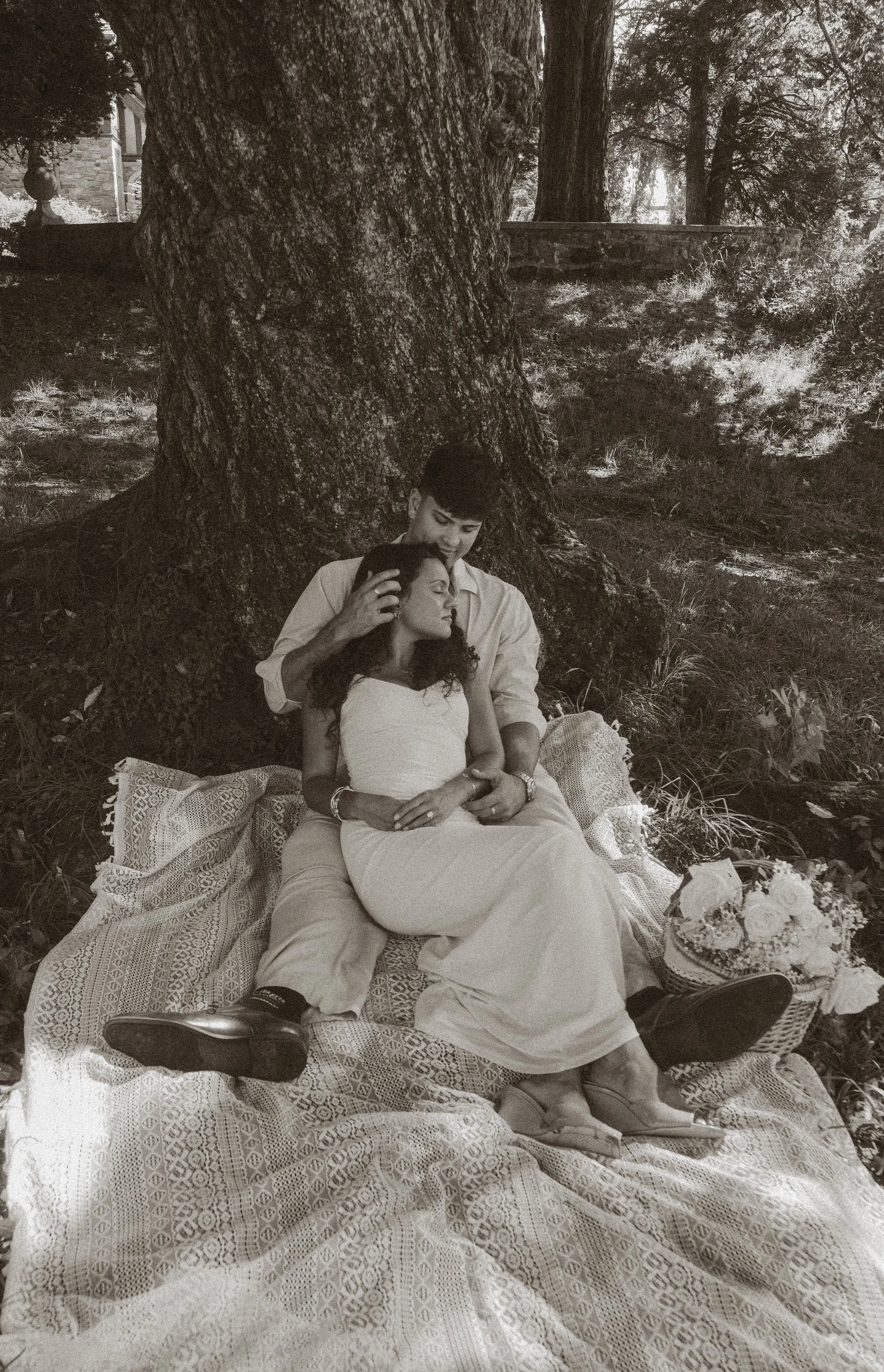 A couple sitting under a large tree on a picnic blanket. The woman is resting with her eyes closed while the man is gently touching her head. There are flowers and a cake nearby.
