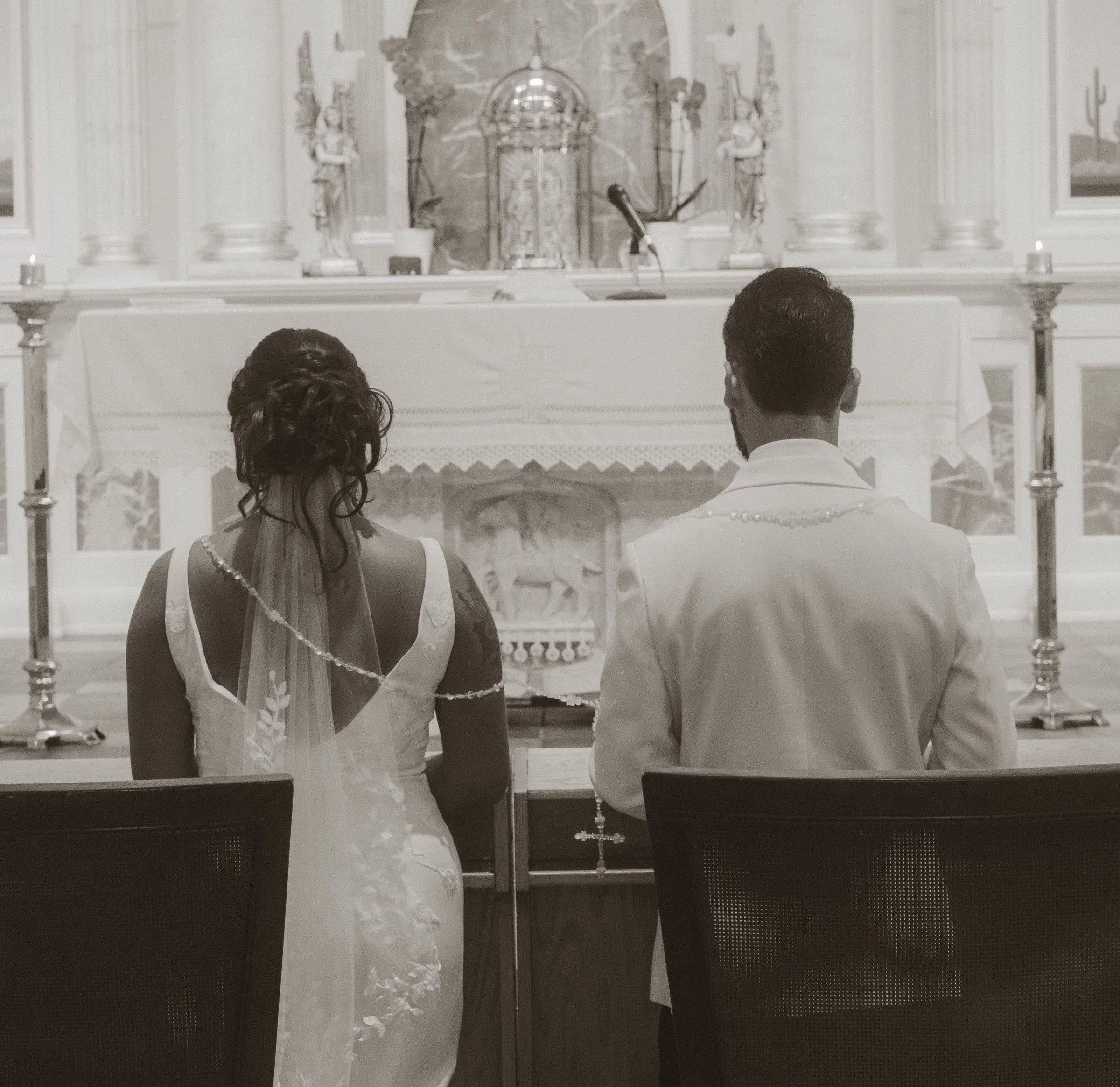 A couple dressed in white sitting in a church pew during a wedding ceremony, seen from behind. The bride has either braided or styled hair, and the groom has short dark hair. The altar behind them features religious statues, a tabernacle, and candles.