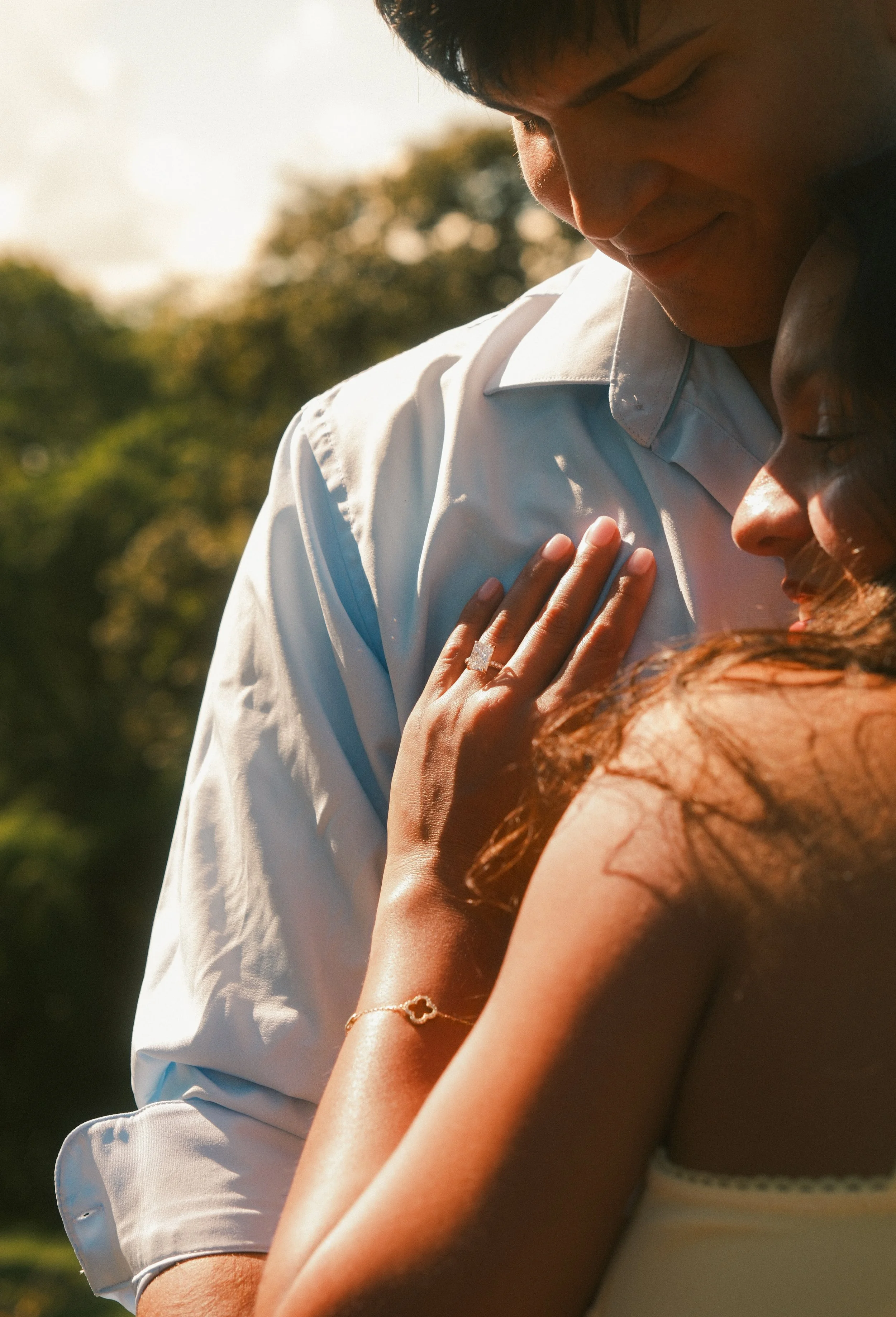 A young man in a white shirt and a woman in a yellow top share a tender moment outdoors, with sunlight and trees in the background.