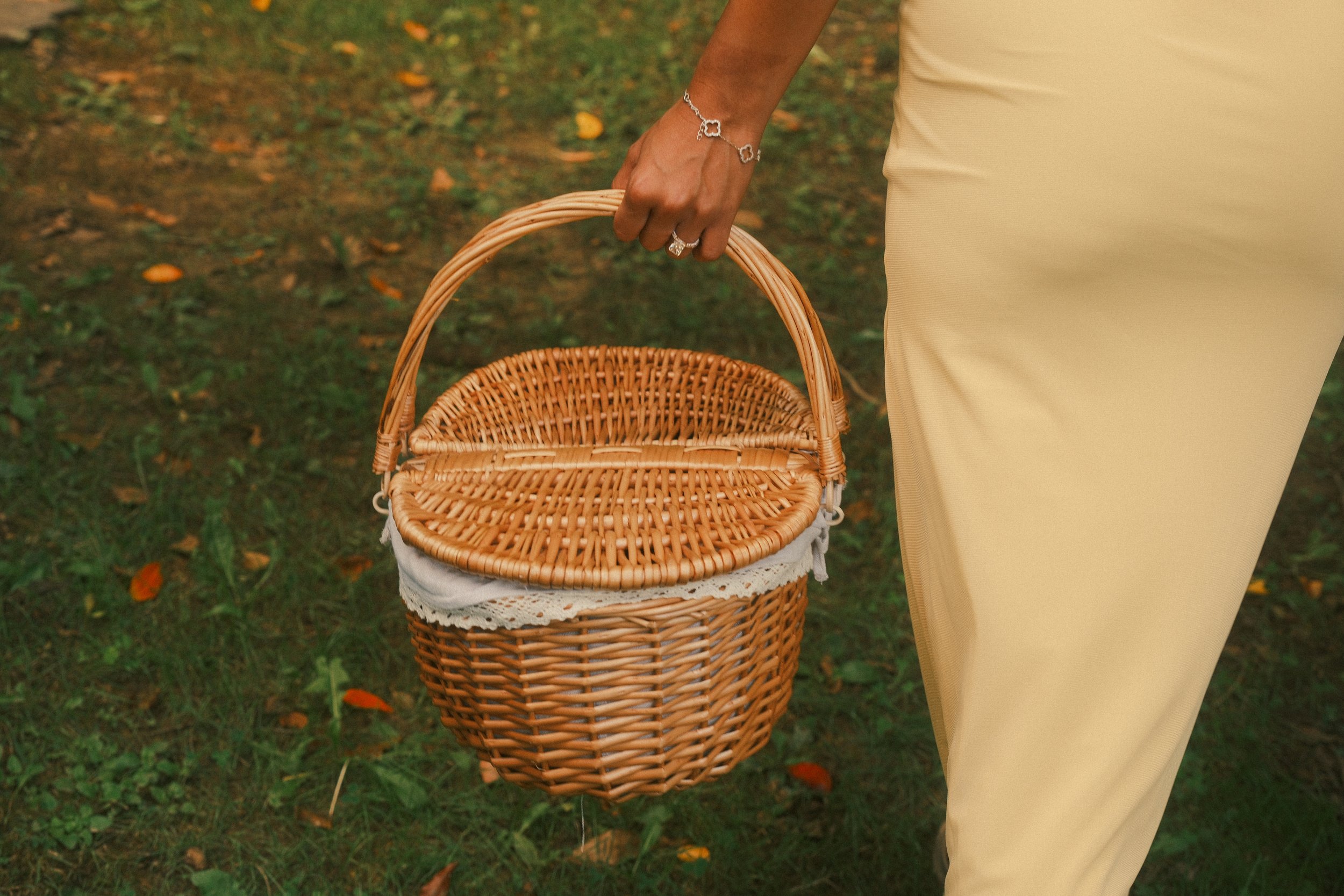 Image of a person's hand holding a wicker basket, standing outdoors on grass with fallen leaves, wearing jewelry including rings and a bracelet, and dressed in beige pants.