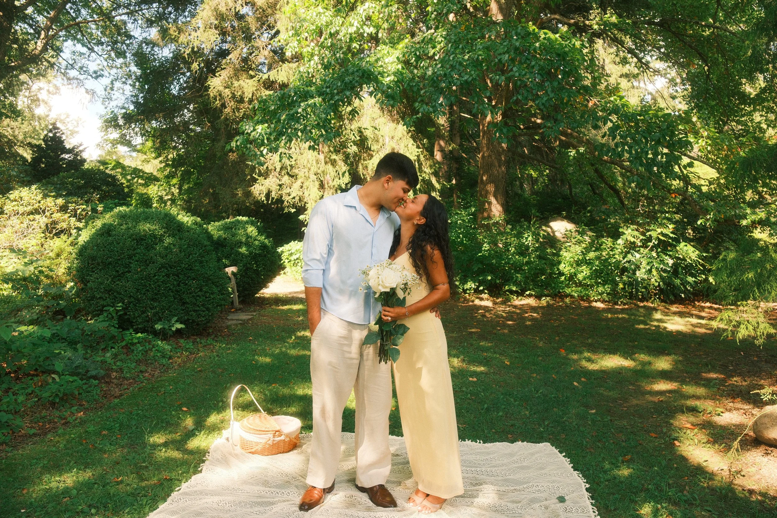 A couple is kissing outdoors in a park, standing on a white rug with a picnic basket nearby. The man is wearing a light blue shirt and beige pants, and the woman is dressed in a cream-colored outfit, holding a bouquet of white roses. Surrounded by lu