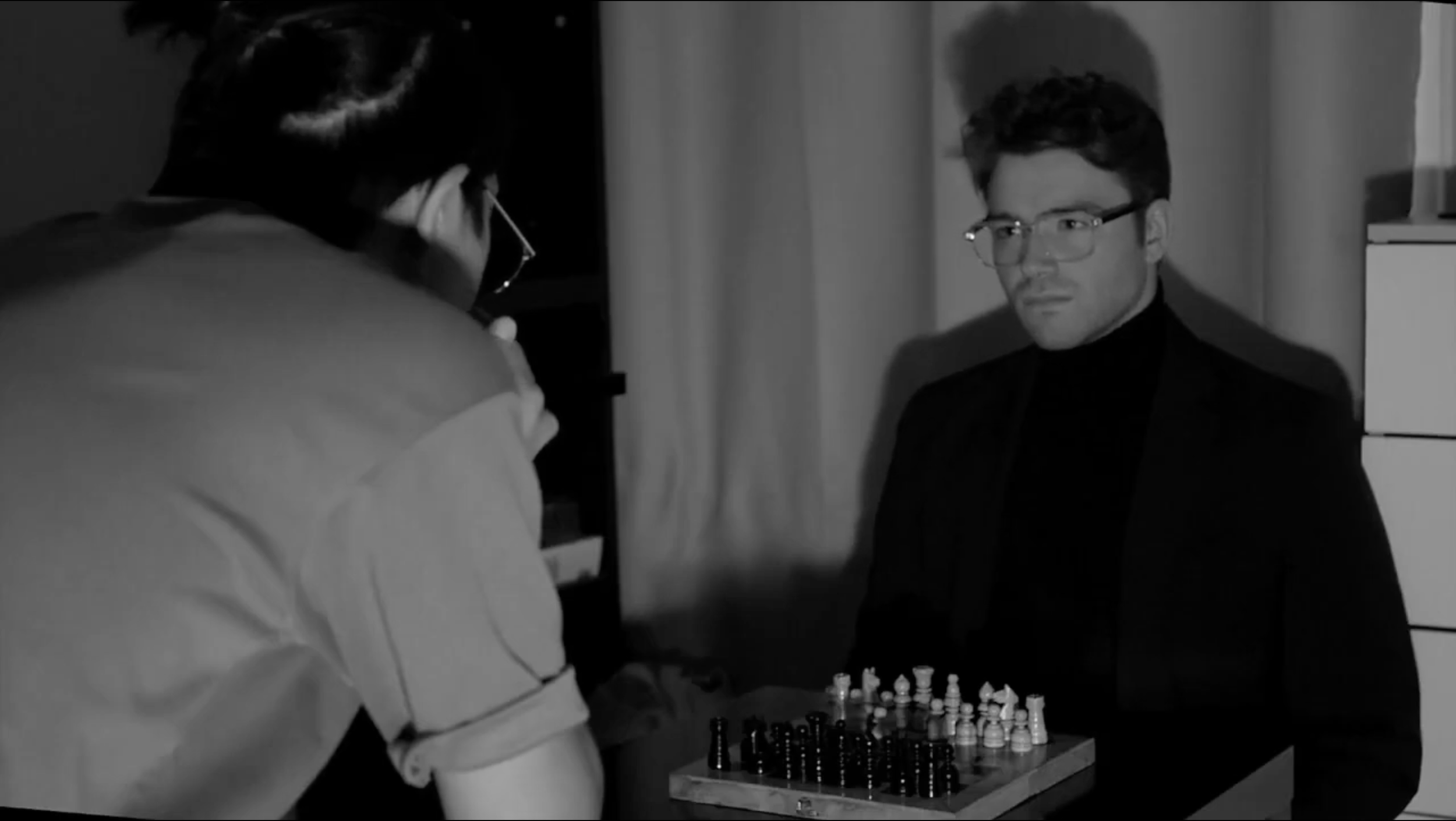 Deux hommes jouent aux échecs dans une pièce sombre, en noir et blanc. L'un, vu de dos, porte une chemise légère, l'autre porte une veste noire avec des lunettes.
