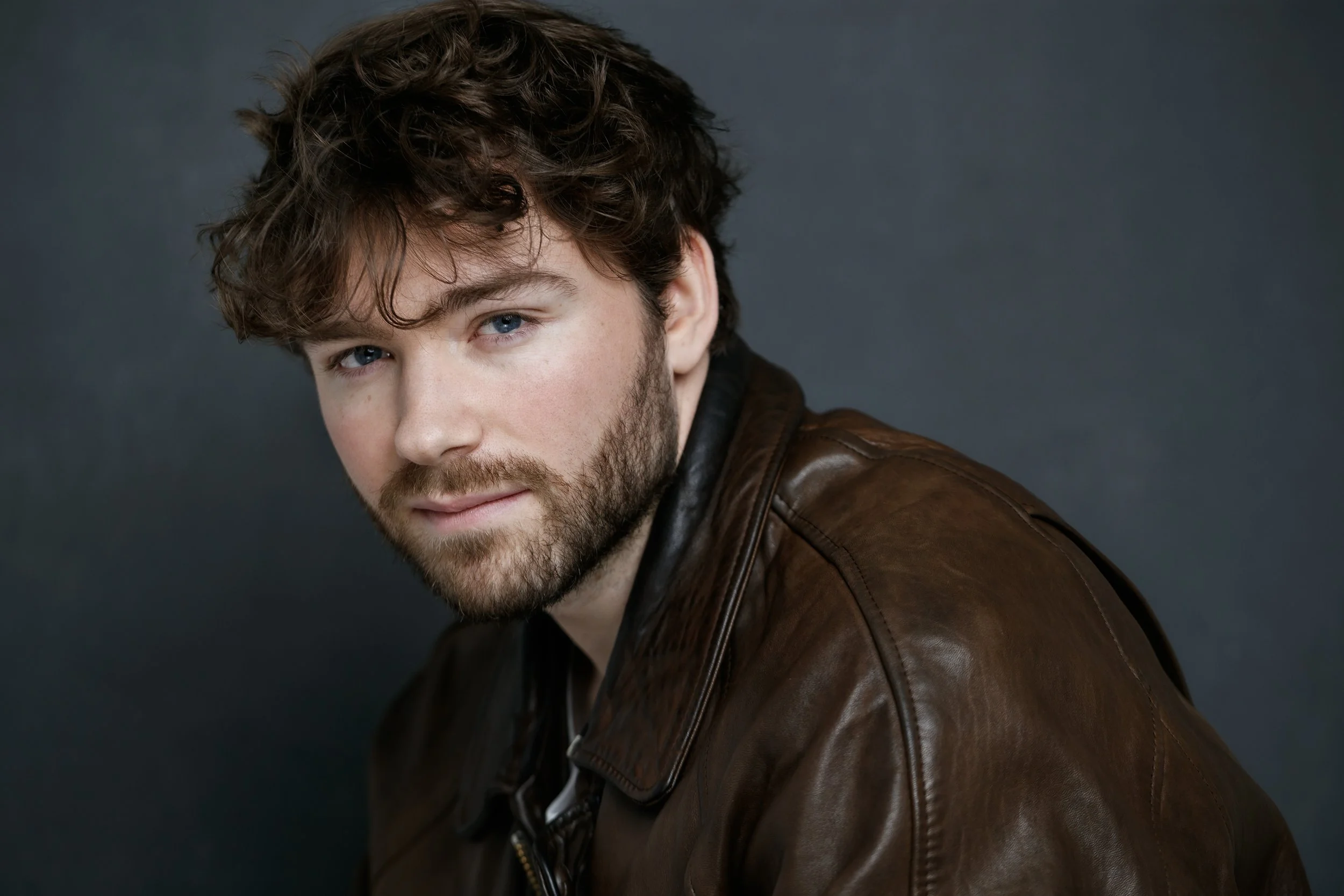 Portrait de Raphaël Gareth portant une veste en cuir marron, contre un fond gris foncé.