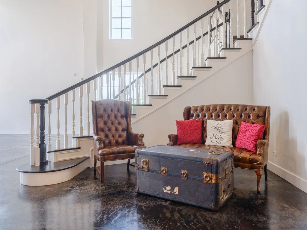 Vintage leather armchair and sofa with decorative pillows, a trunk, and a staircase in a bright, modern living room.