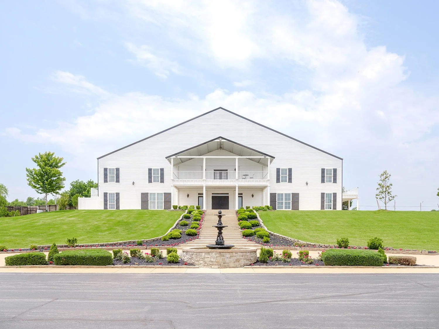 Large white two-story house with black shutters, front porch, and landscaped lawn with bushes, flowers, and a fountain, under a blue sky.