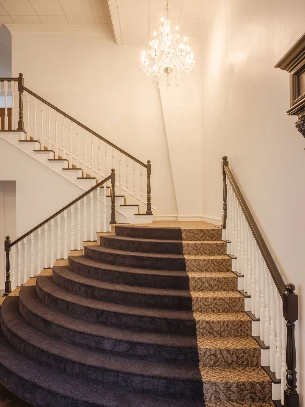 A staircase with dark carpeted steps transitions into a light-colored carpeted area at the top. The staircase has white balusters and dark handrails. A crystal chandelier hangs from the ceiling, illuminating the area.