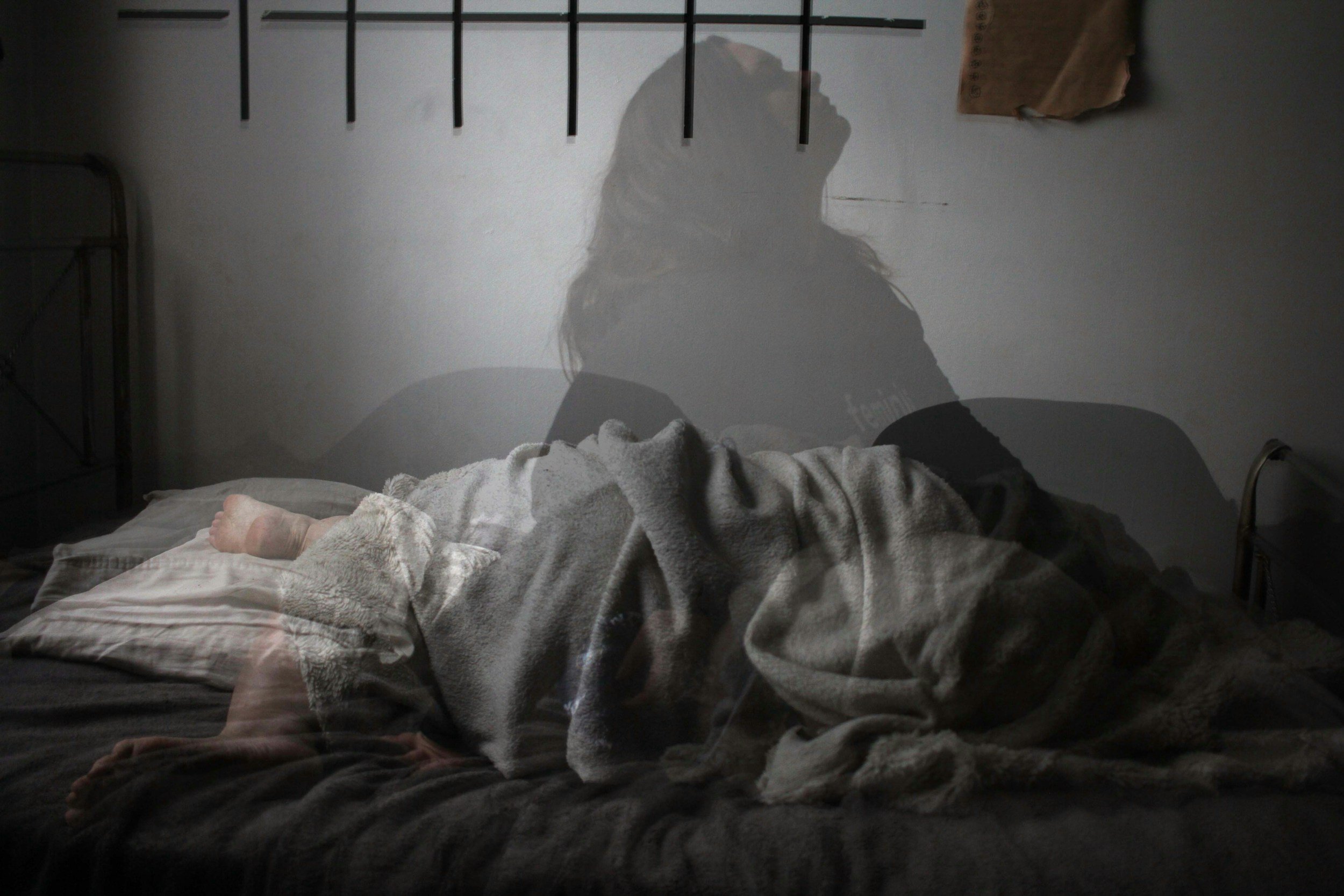 A person lying on a bed wrapped in a blanket, with their head on a pillow, captured through a window creating a double exposure effect with their silhouette visible against a plain wall.