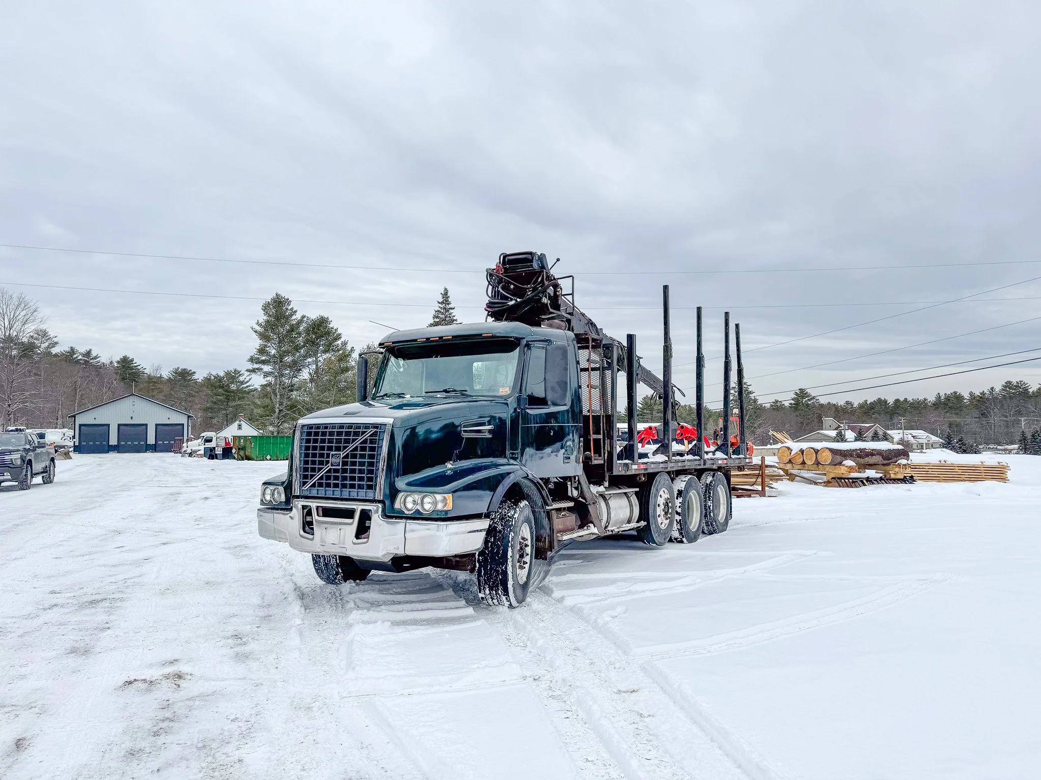 A black logging truck with a crane on top parked on snow-covered ground in a rural area with trees and buildings in the background. Tree Service Maine, Learned Tree Service 