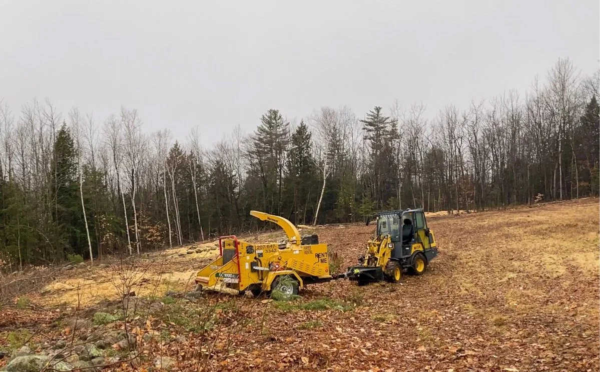 A small yellow wood chipper being operated by a person on a yellow compact utility loader in a clearing surrounded by trees with autumn foliage. Tree Service Maine, Learned Tree Service 