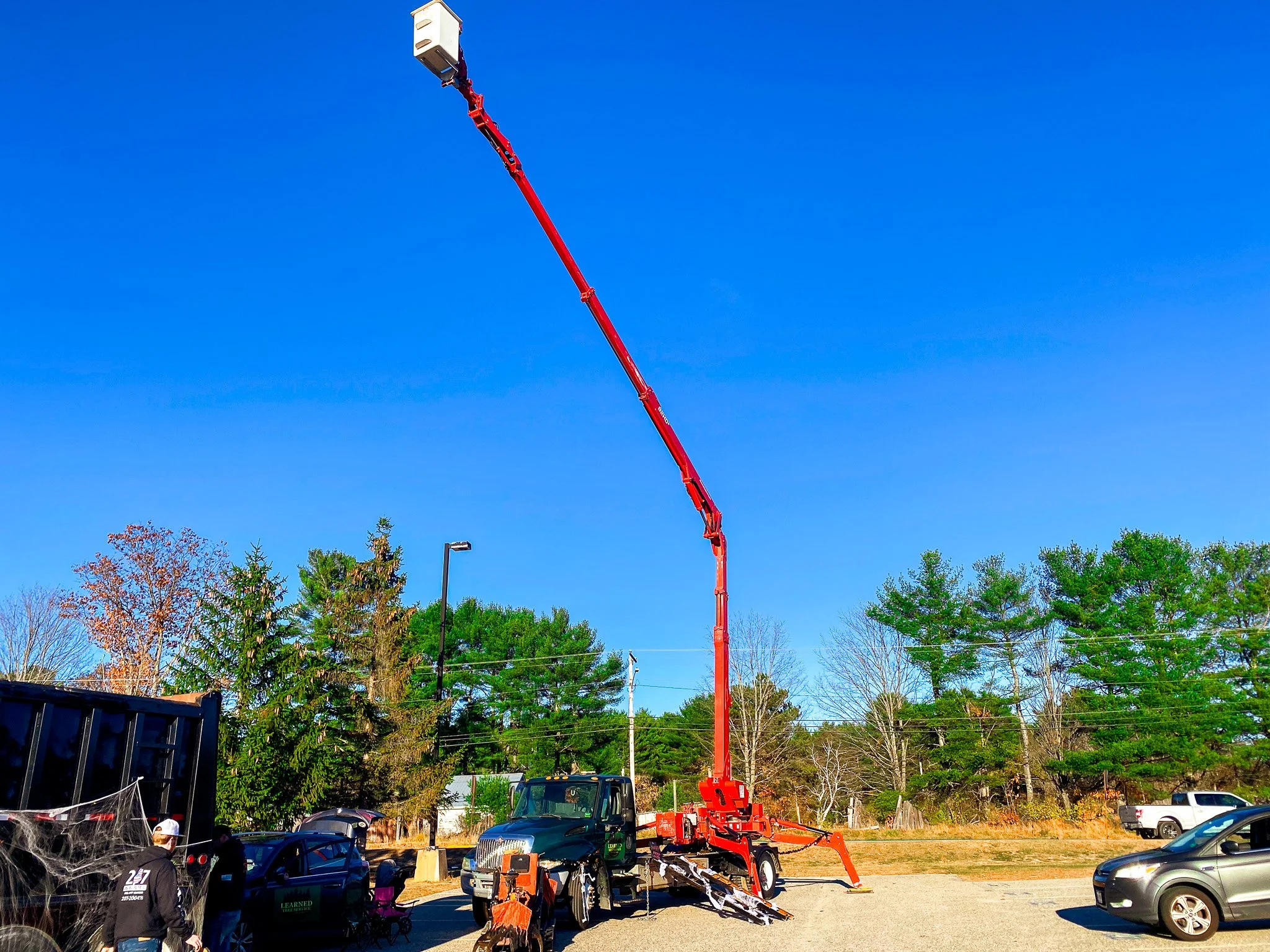 A large cherry picker with a long red arm extended upward, parked in a gravel lot with cars and trucks around, and trees and a clear blue sky in the background. Tree Service Maine, Learned Tree Service 