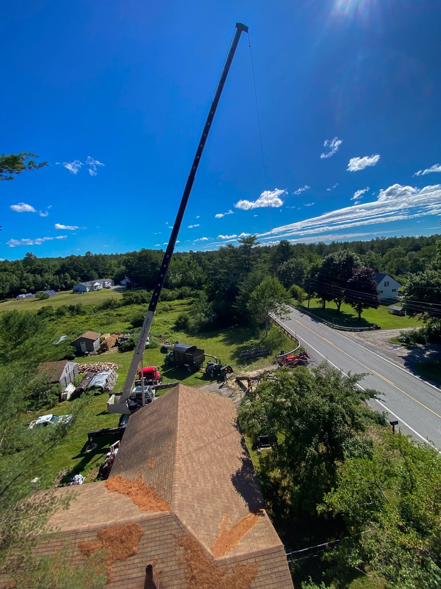 A construction crane is lifting roofing materials onto a house under a bright blue sky with scattered clouds. The house has a brown shingle roof, and there are trees and a road nearby. Tree Service Maine, Learned Tree Service