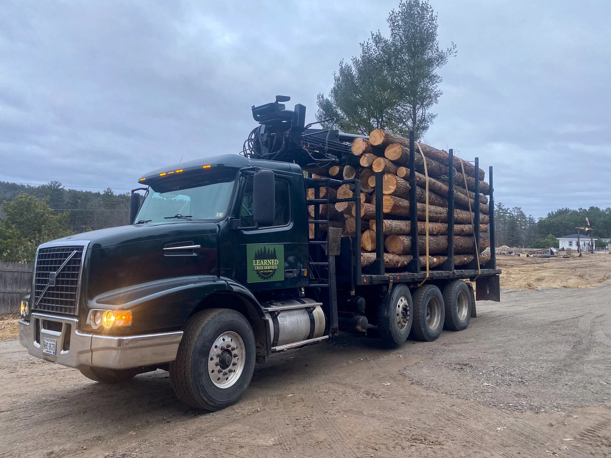 A black logging truck carrying cut logs on a dirt lot with a cloudy sky in the background. Tree Service Maine, Learned Tree Service 