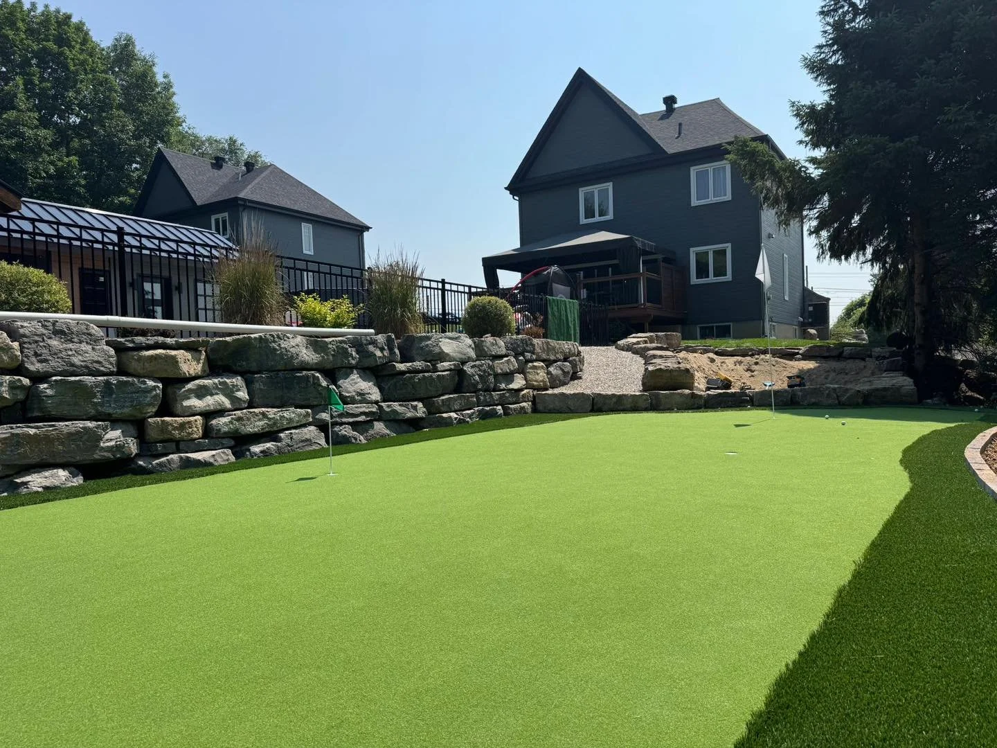 Terrain de mini-golf avec des trous, un mur en pierre et une maison en arrière-plan, en plein jour.