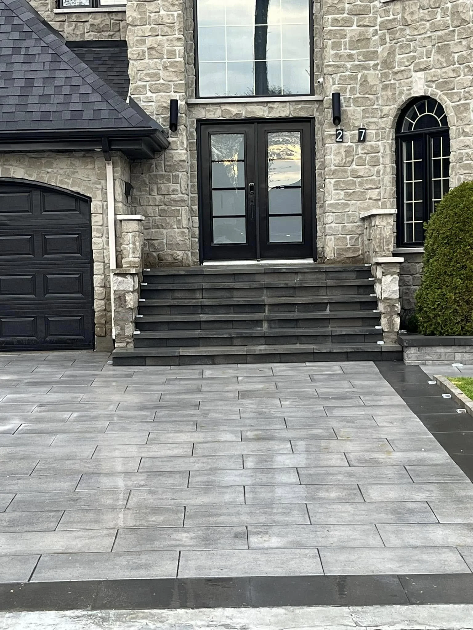 Façade d'une maison en pierre avec escalier noir menant à une porte d'entrée vitrée noire, entourée de murs en pierre, avec un garage à gauche et un buisson à droite.