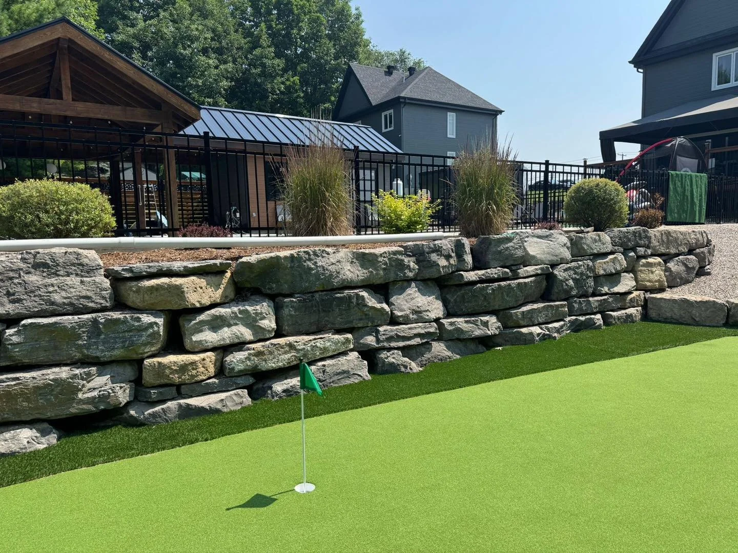 Terrain de mini-golf avec un trou et un drapeau vert, mur en pierre derrière une terrasse avec mobilier, arbres, maisons en arrière-plan, ciel clair.