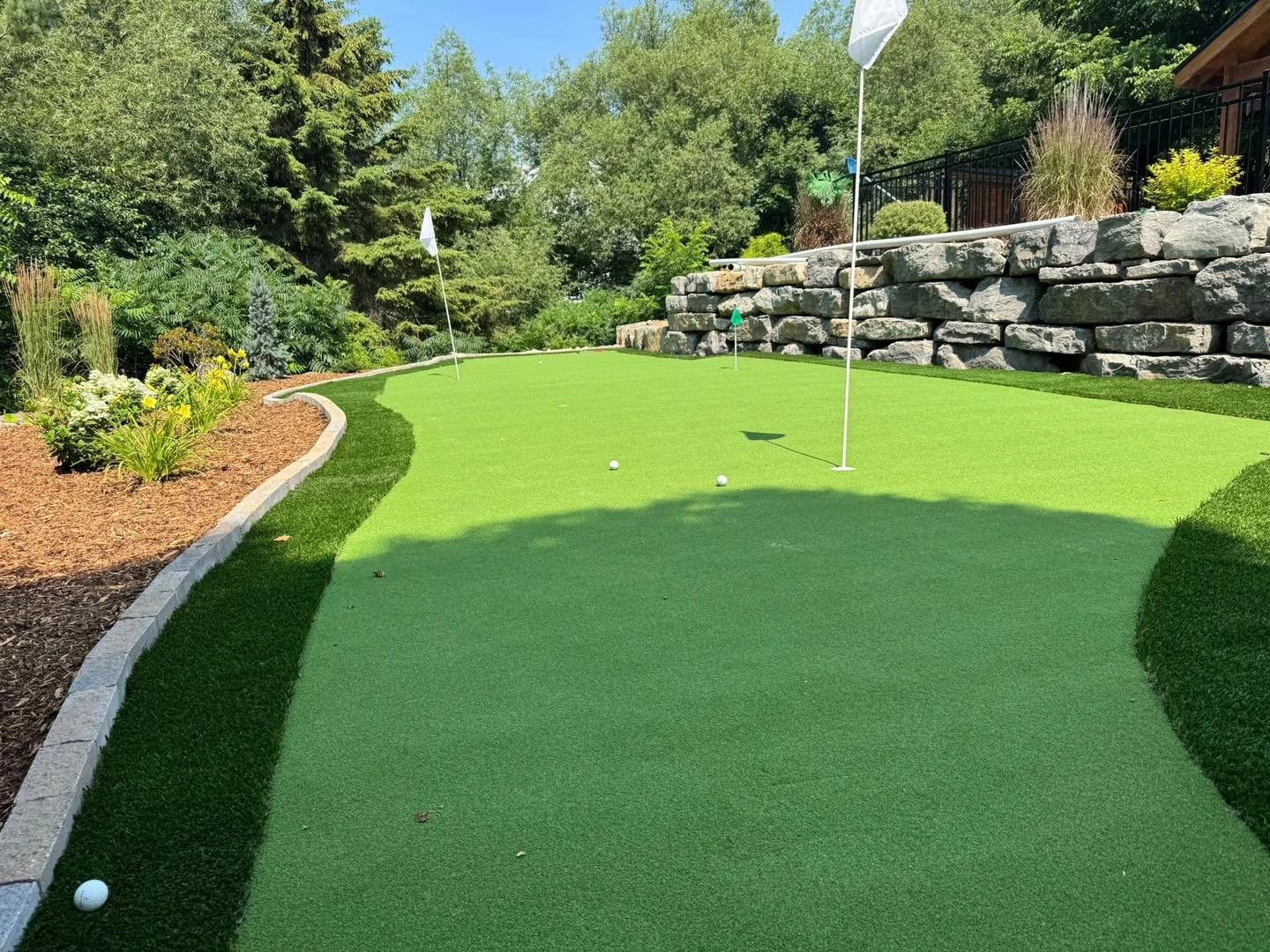 Un mini parcours de golf avec trois balles de golf et trois drapeaux blancs, entouré de verdure, de fleurs et d'un mur en pierre.