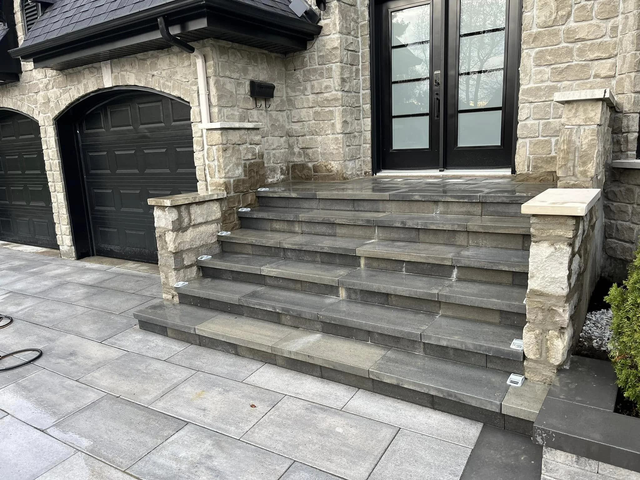 Escaliers en pierre menant à une porte d'entrée noire dans une maison en pierre, avec deux garages noirs sur le côté gauche et un sol en pavés gris.