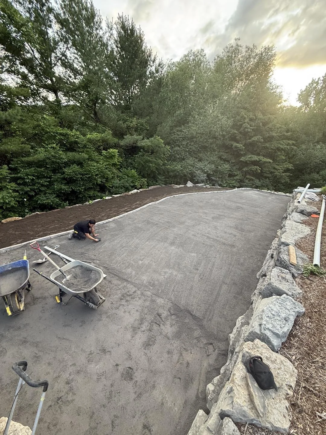 Une personne travaille à créer une surface de béton dans un jardin entouré d'arbres, avec des outils comme une brouette et une pelle, lors d'une journée ensoleillée.