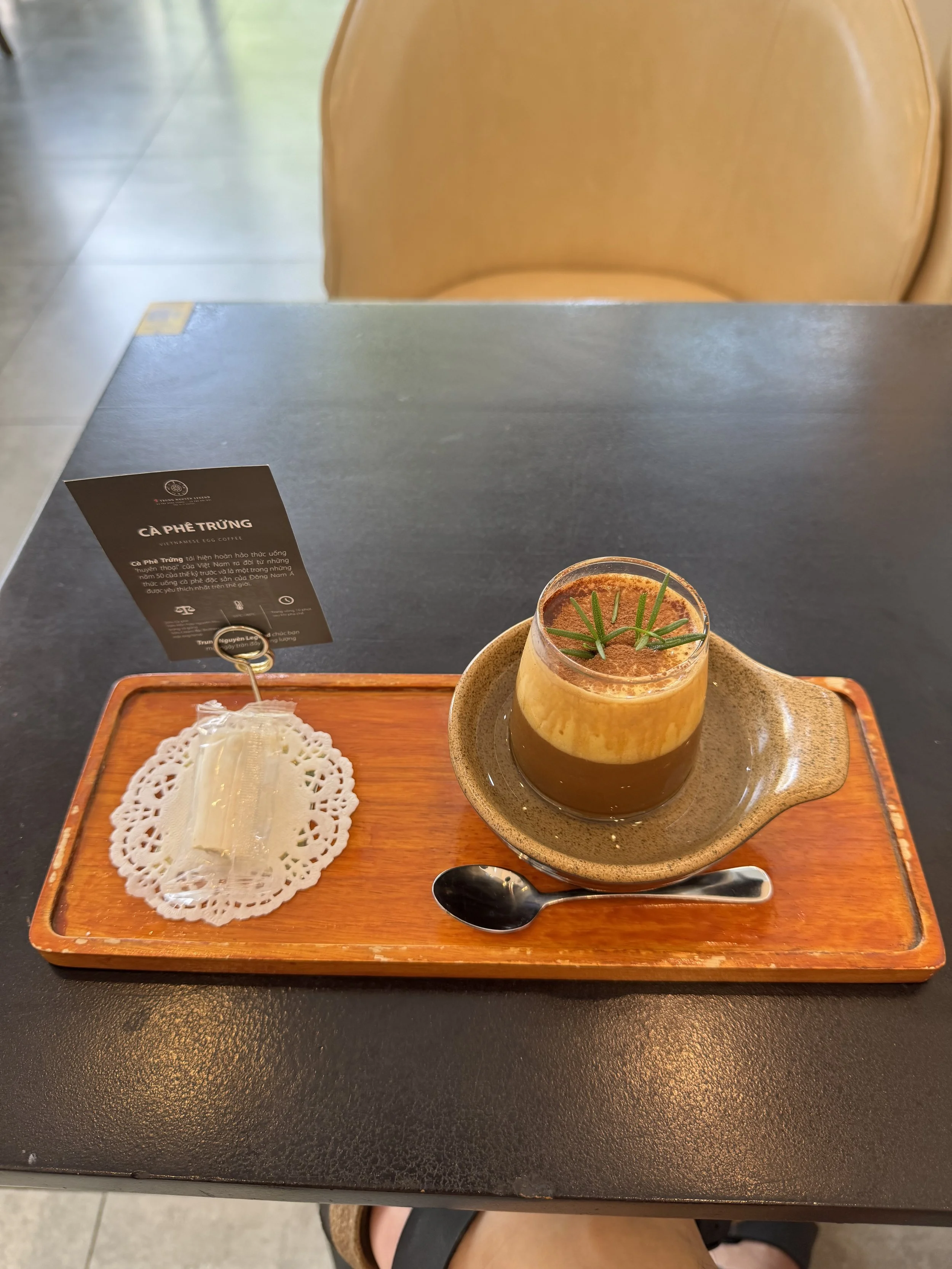 A glass of Vietnamese egg coffee with a sprig of rosemary on top, served on a wooden tray with a small spoon and a wrapped piece of sugar, in a café setting.