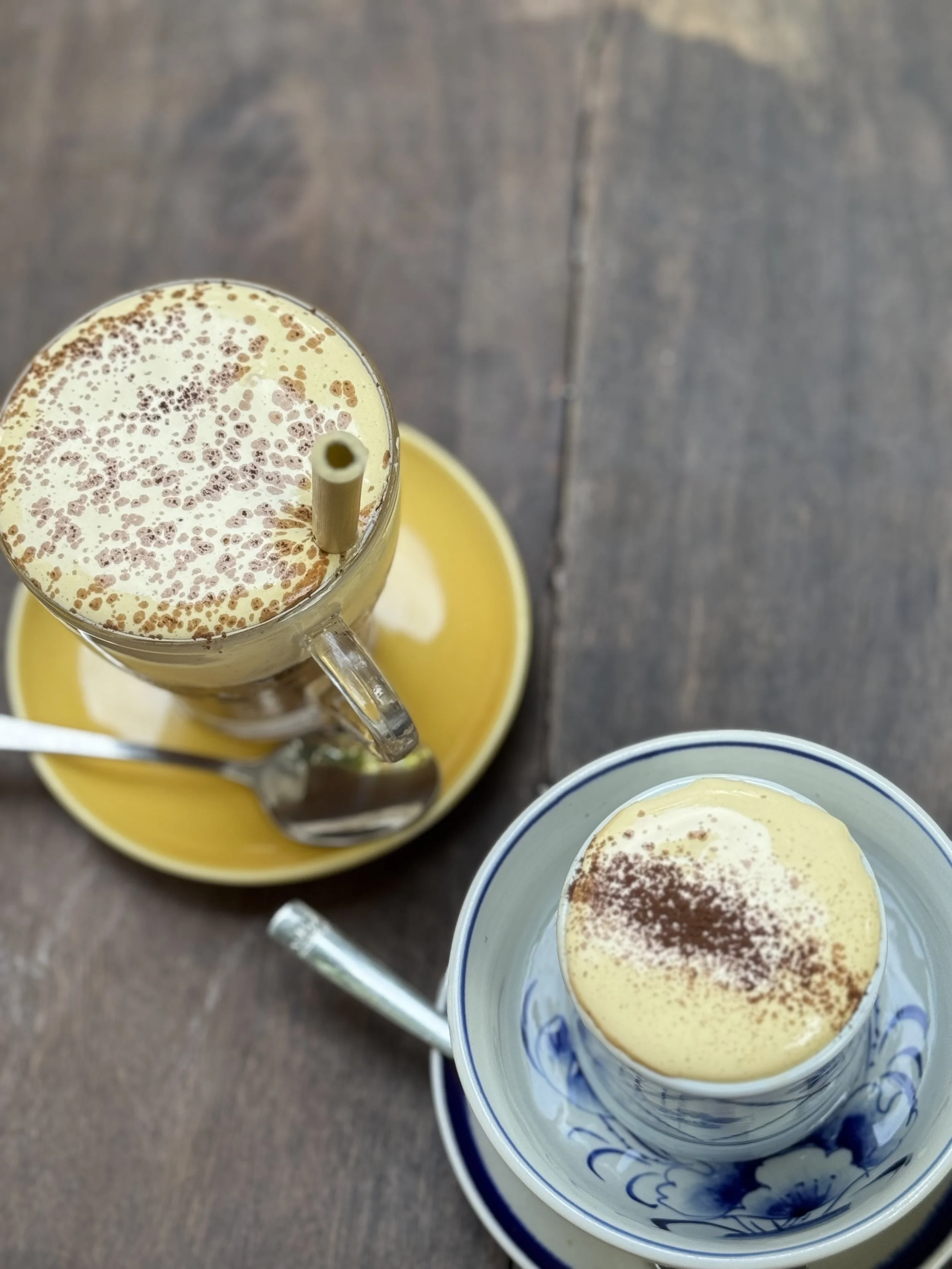 Two glasses of yellow-colored beverages topped with frothy cream and sprinkled with cocoa powder, served on small saucers with spoons, on a wooden table.