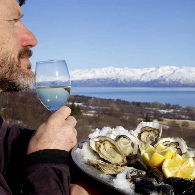 A man holding a glass of white wine, with a plate of fresh oysters and lemon wedges in front of him, overlooking a body of water with snow-capped mountains in the background.