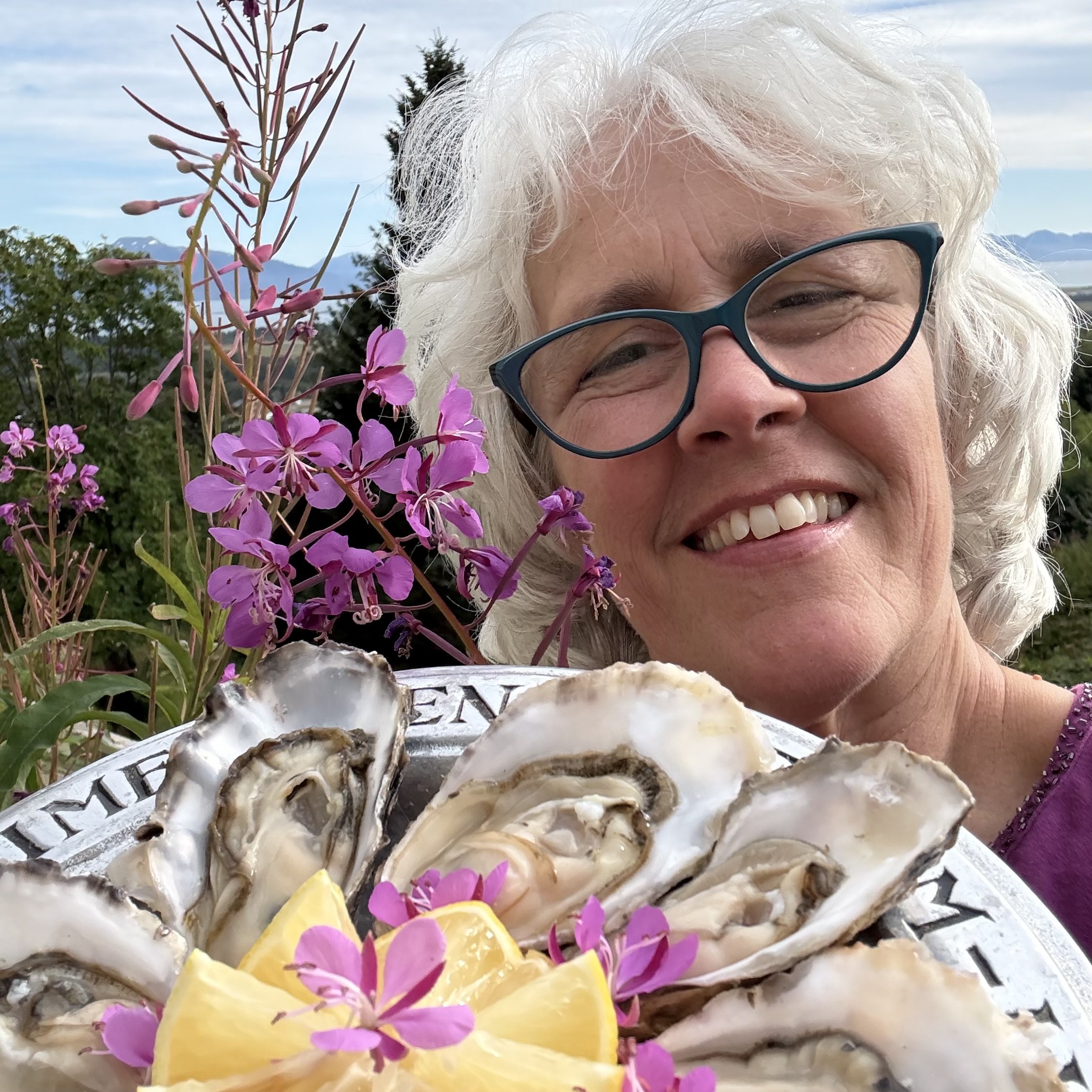 Smiling woman with white hair and glasses outdoors holding a plate of oysters garnished with lemon and fireweed flowers.