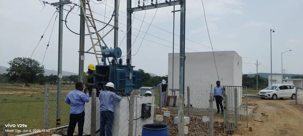 Workers inspecting electrical transformer near power poles in a rural area, with some people in safety helmets, a white building, and a white vehicle in the background.