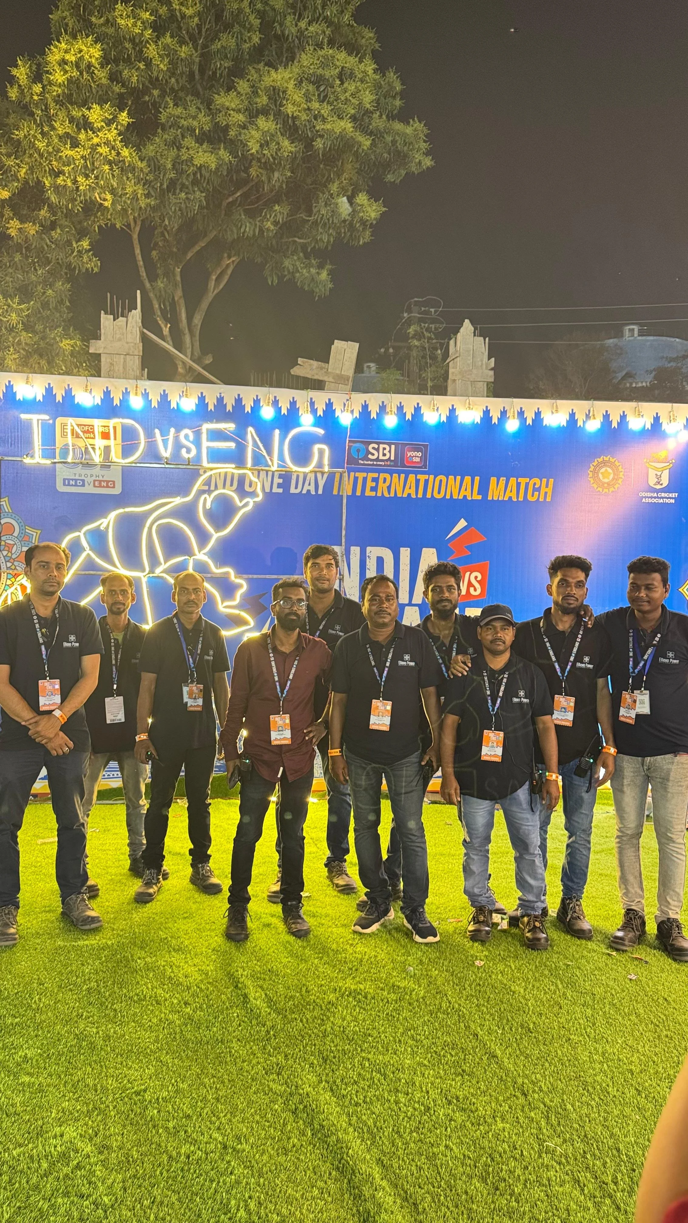 Group of nine men standing on a grass field in front of a blue banner with text that reads "INDIA vs ENG" and mentions the tournament and match details. The men are posing for a photo at night, some with arms around each other, all wearing black shirts with event badges around their necks. There is a large tree and some construction materials and lights visible in the background.