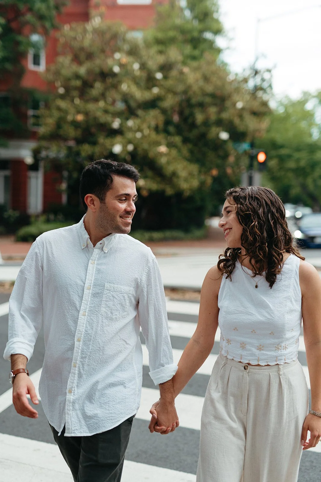 A young couple holding hands and smiling at each other while walking across a crosswalk in a city neighborhood with trees and buildings in the background.
