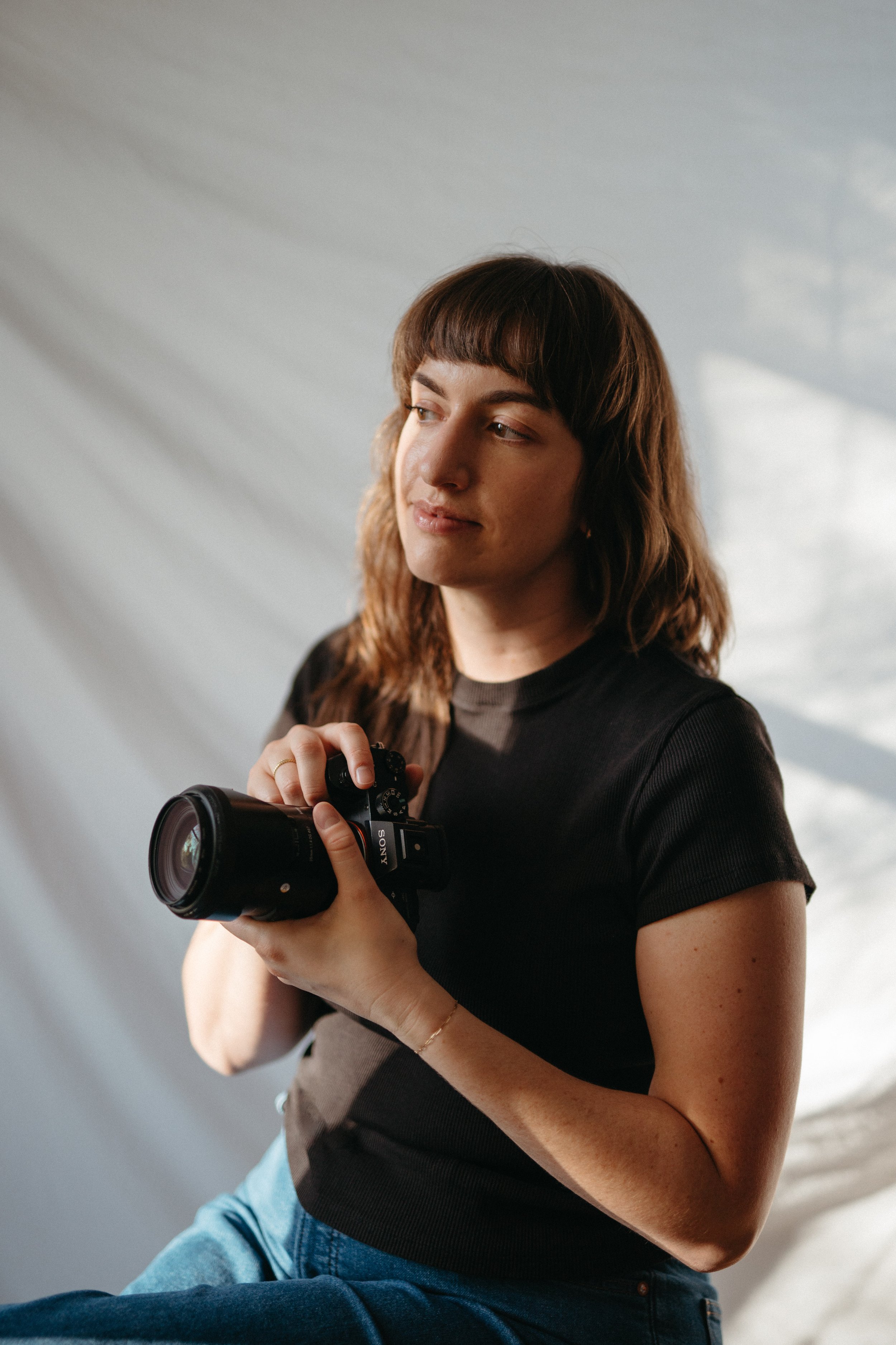 A woman with brown hair and bangs holding a black Sony camera, sitting against a light-colored background.