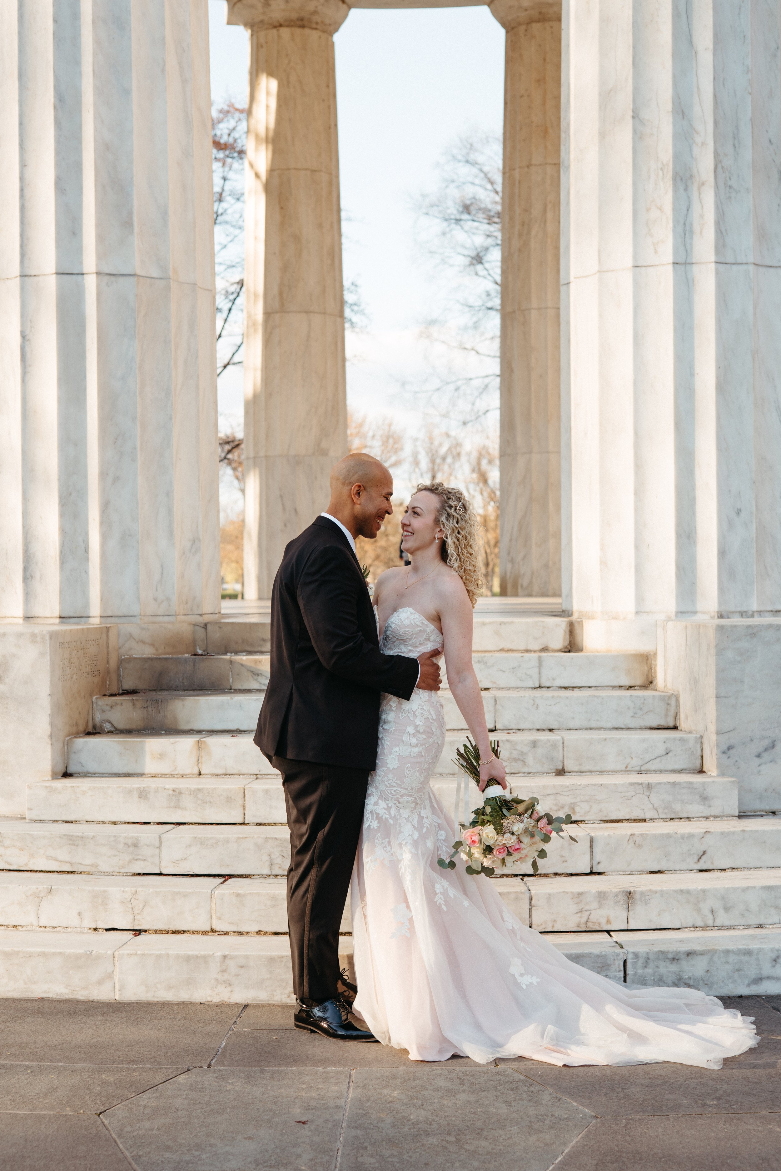 A newlywed couple in wedding attire standing close and smiling at each other in front of marble columns and steps, with the bride holding a bouquet of pink and white flowers.