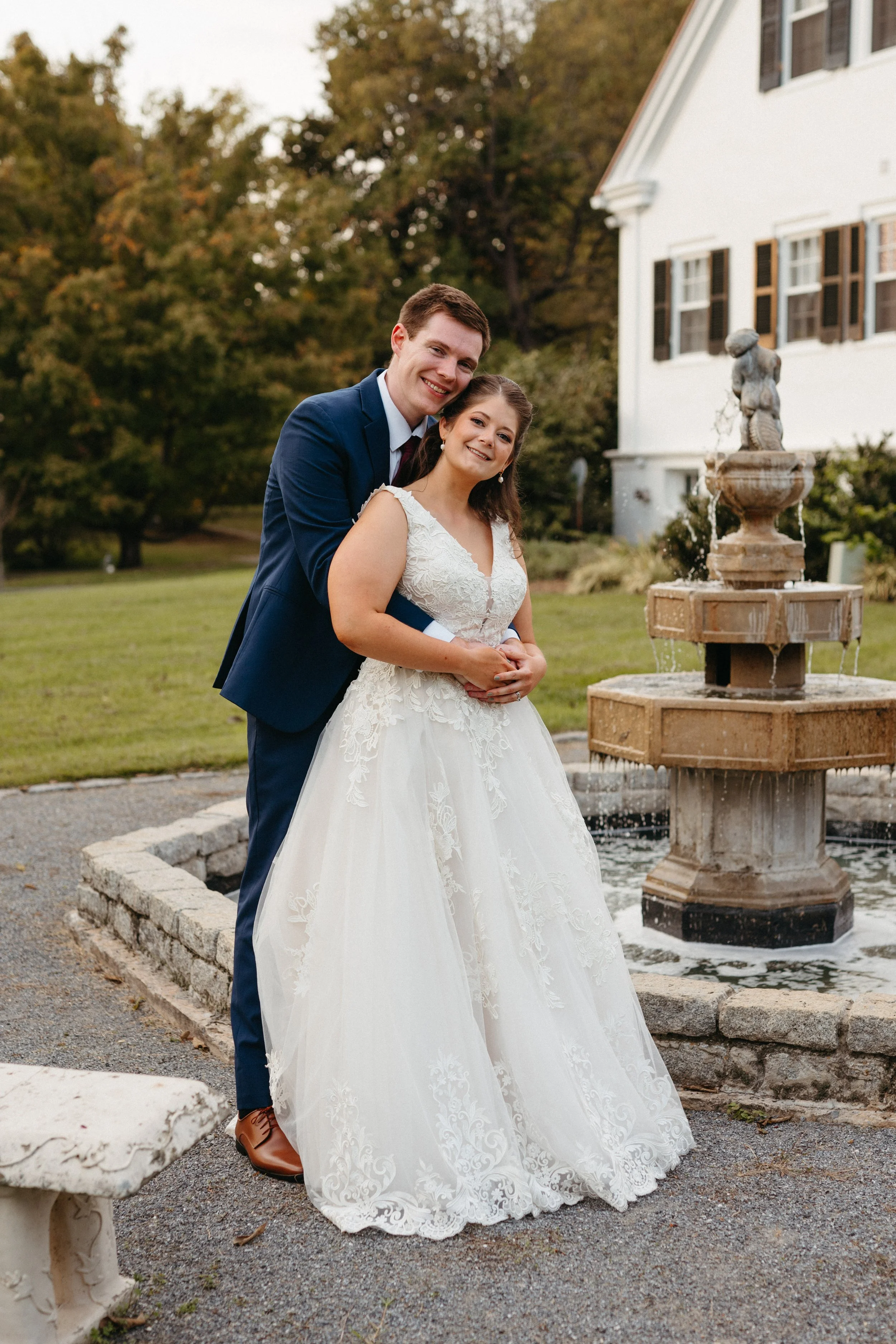 A newlywed couple standing near a fountain outside, with the groom in a navy suit and the bride in a white lace wedding gown, smiling and embracing during sunset.