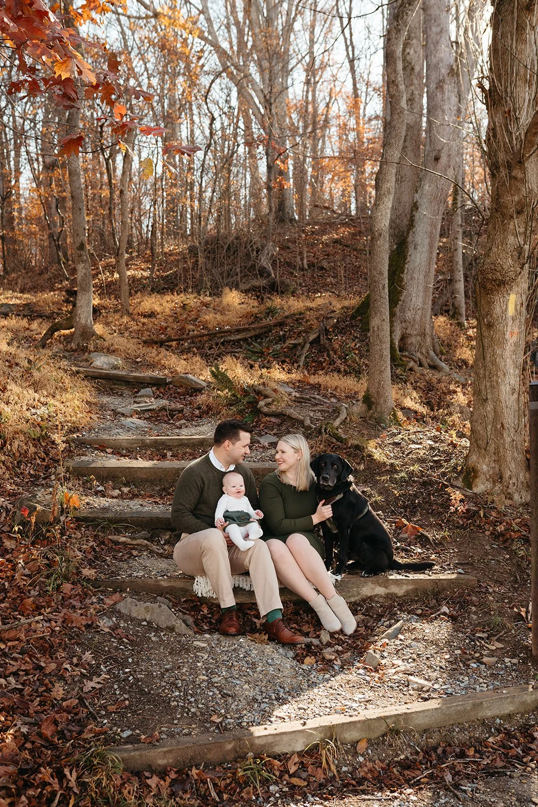 A family of three with a dog sitting on outdoor stairs in a wooded area during fall. The man holds a baby, and the woman is sitting next to a sleeping black dog, all smiling.