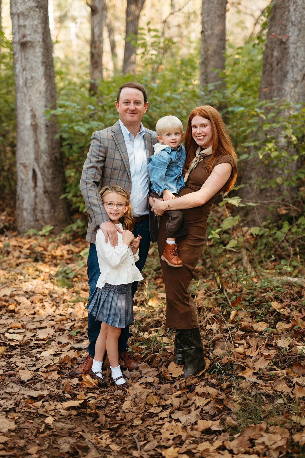 Family of four, two children and two adults, standing on a leaf-covered forest floor during autumn.